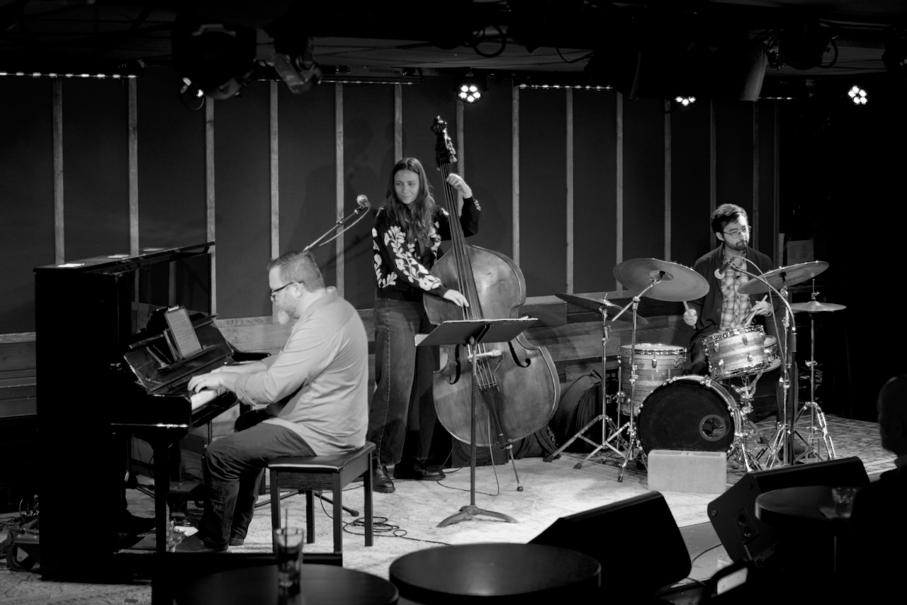 Four musicians perform on stage: a pianist, a double bassist, a drummer, and a vocalist, with a piano and microphones.