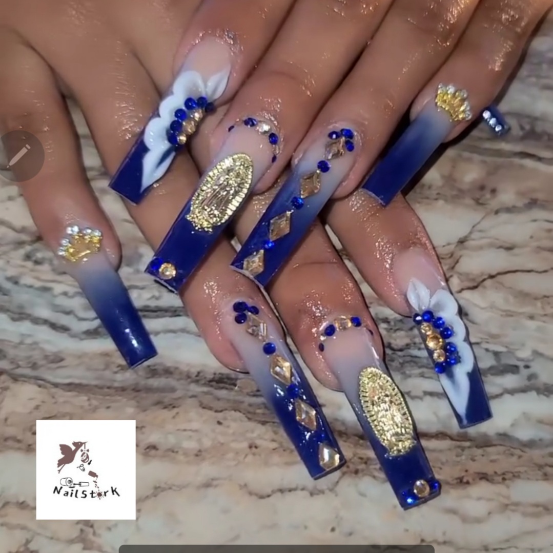 Nail art with blue, gold, and white designs, including flowers, rhinestones, and glitter, on long, decorated nails.