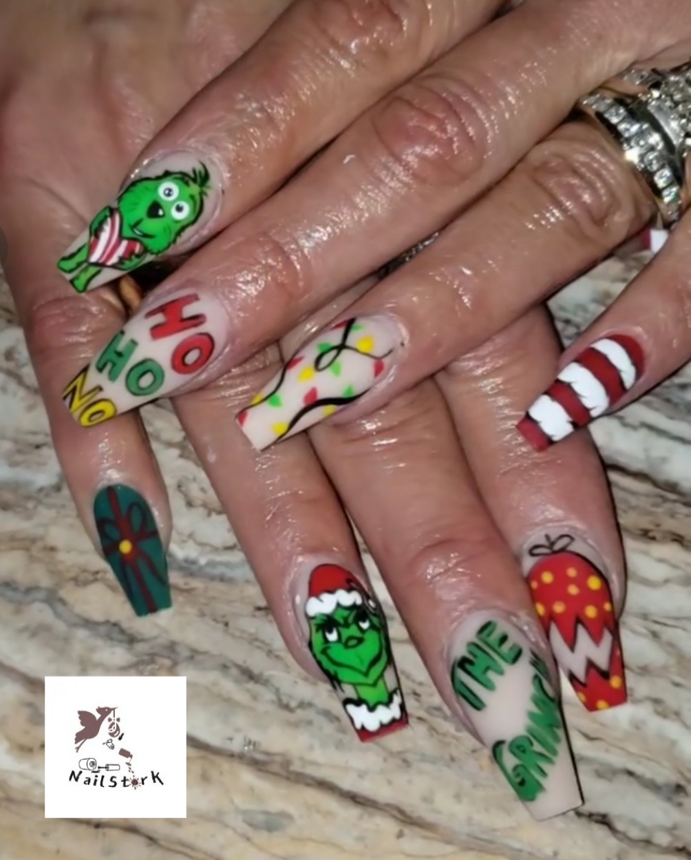 Hands with Christmas-themed nail art, including a fish, a green creature, and holiday patterns, on a marble surface.