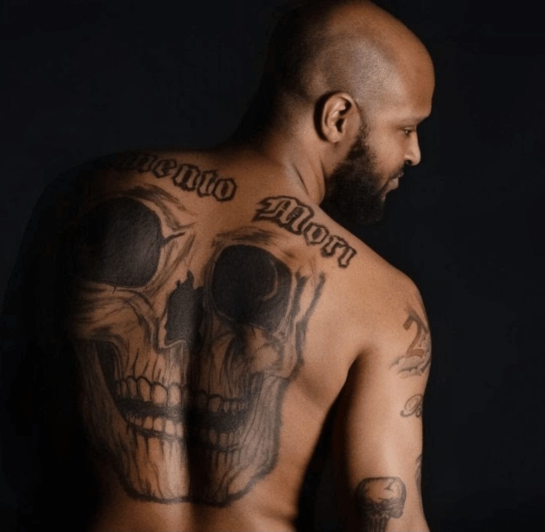 Back view of a shirtless man with skull tattoos on his back and arm, facing right, dark background.
