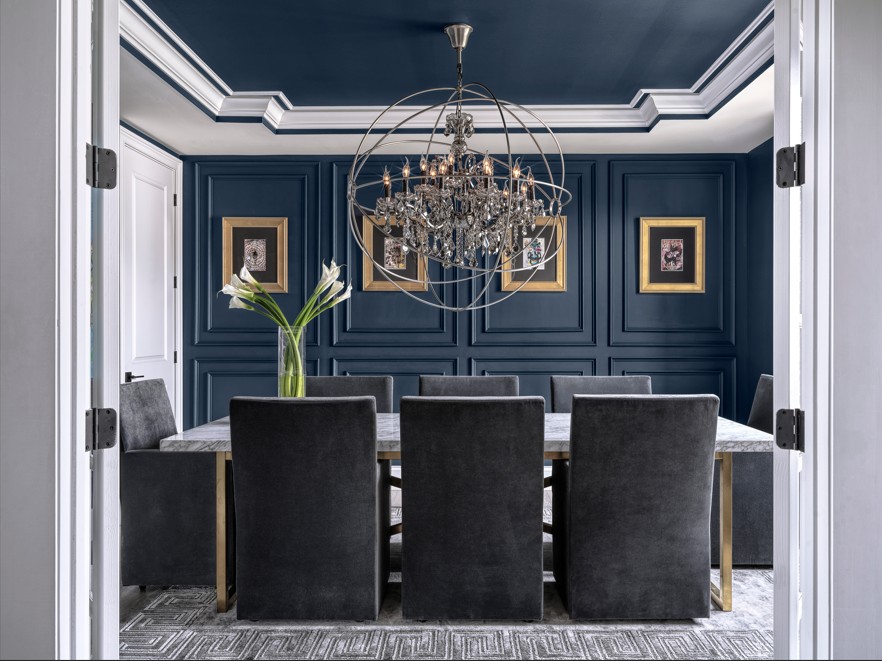 Dining room with a chandelier, blue paneled walls, a table with six chairs, and a vase with white flowers.