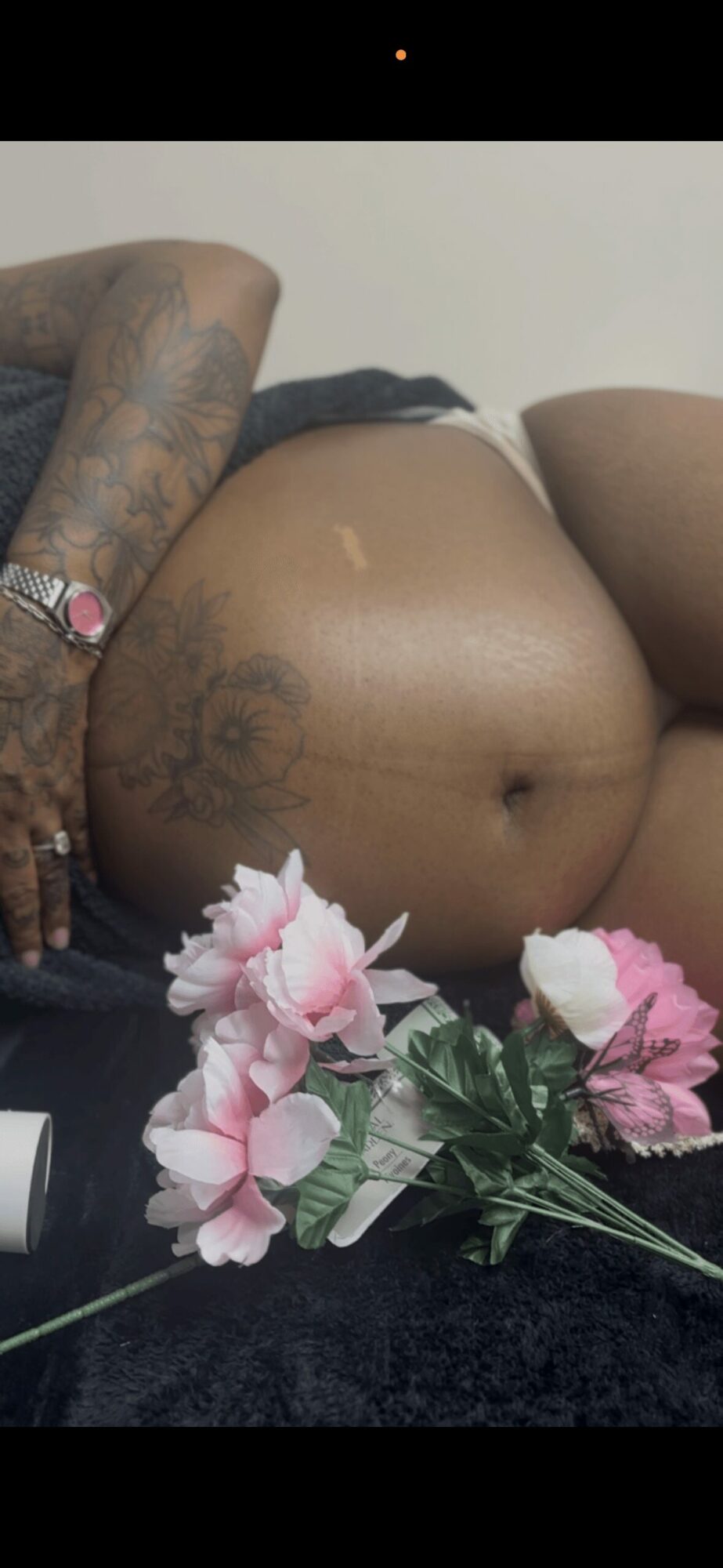 Close-up of a person's back and buttocks with tattoos, flowers in foreground, and a plain background.