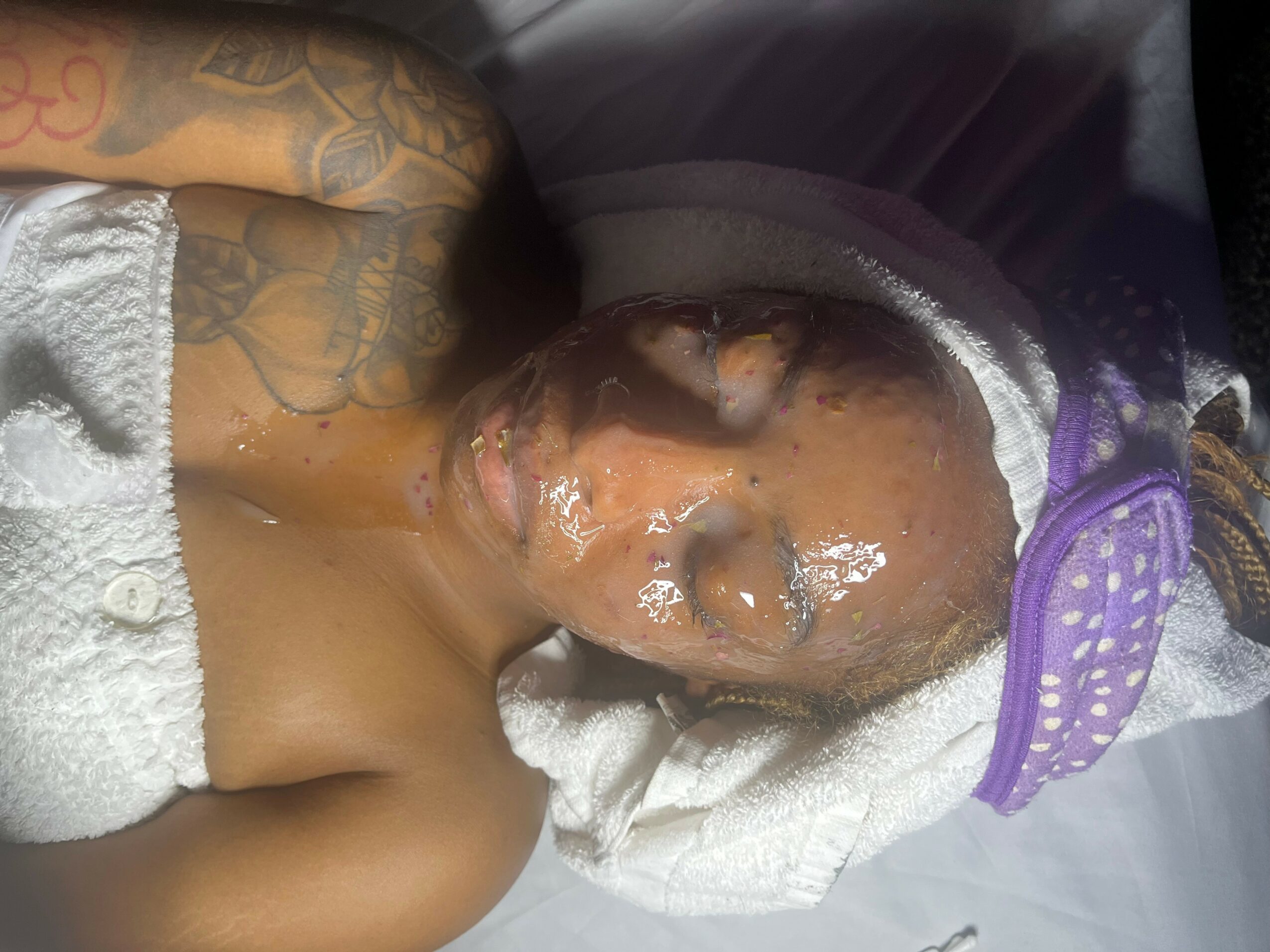 A person with tattoos lying on a hospital bed with a towel on their chest and a towel wrapped around their head, eyes closed.