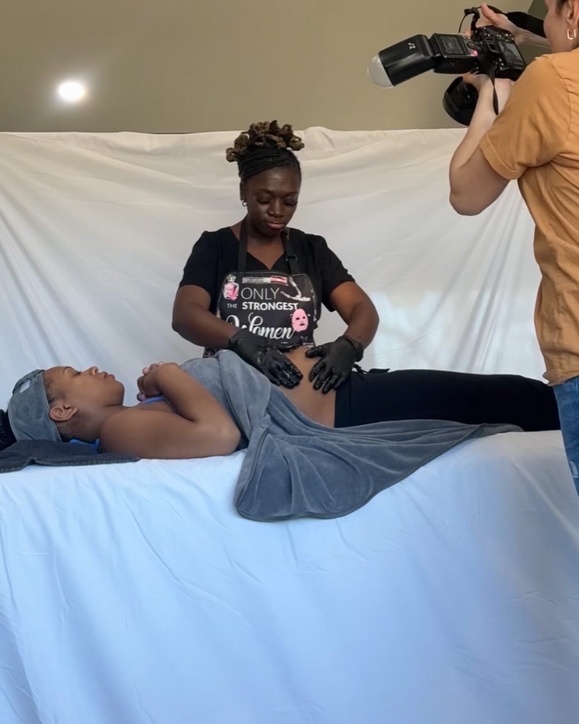 Person lying on a table with a towel, being filmed by a person with a camera, in a studio setting.