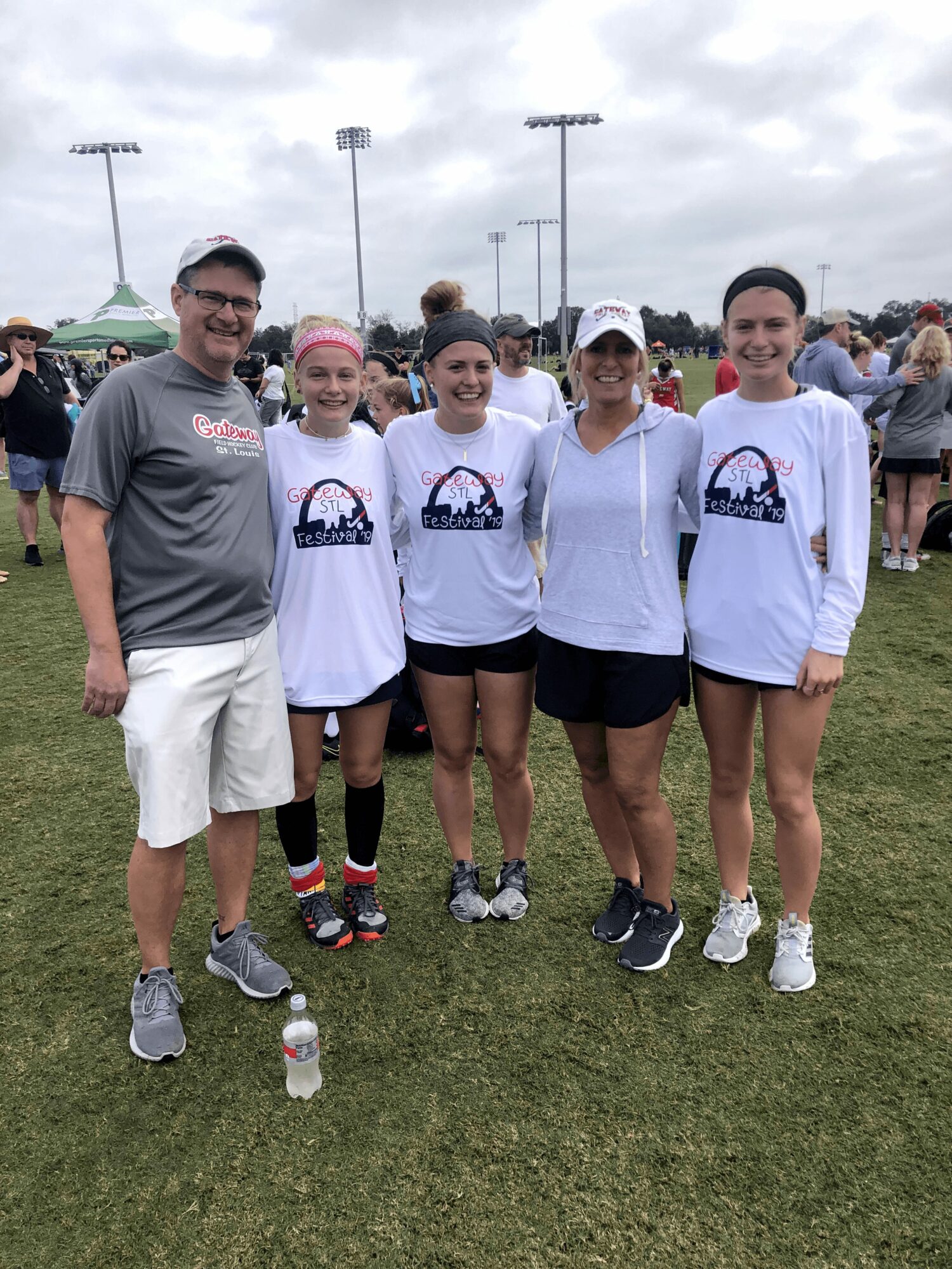 Group of five people standing outdoors on grass, smiling, with a sports field and cloudy sky in background.
