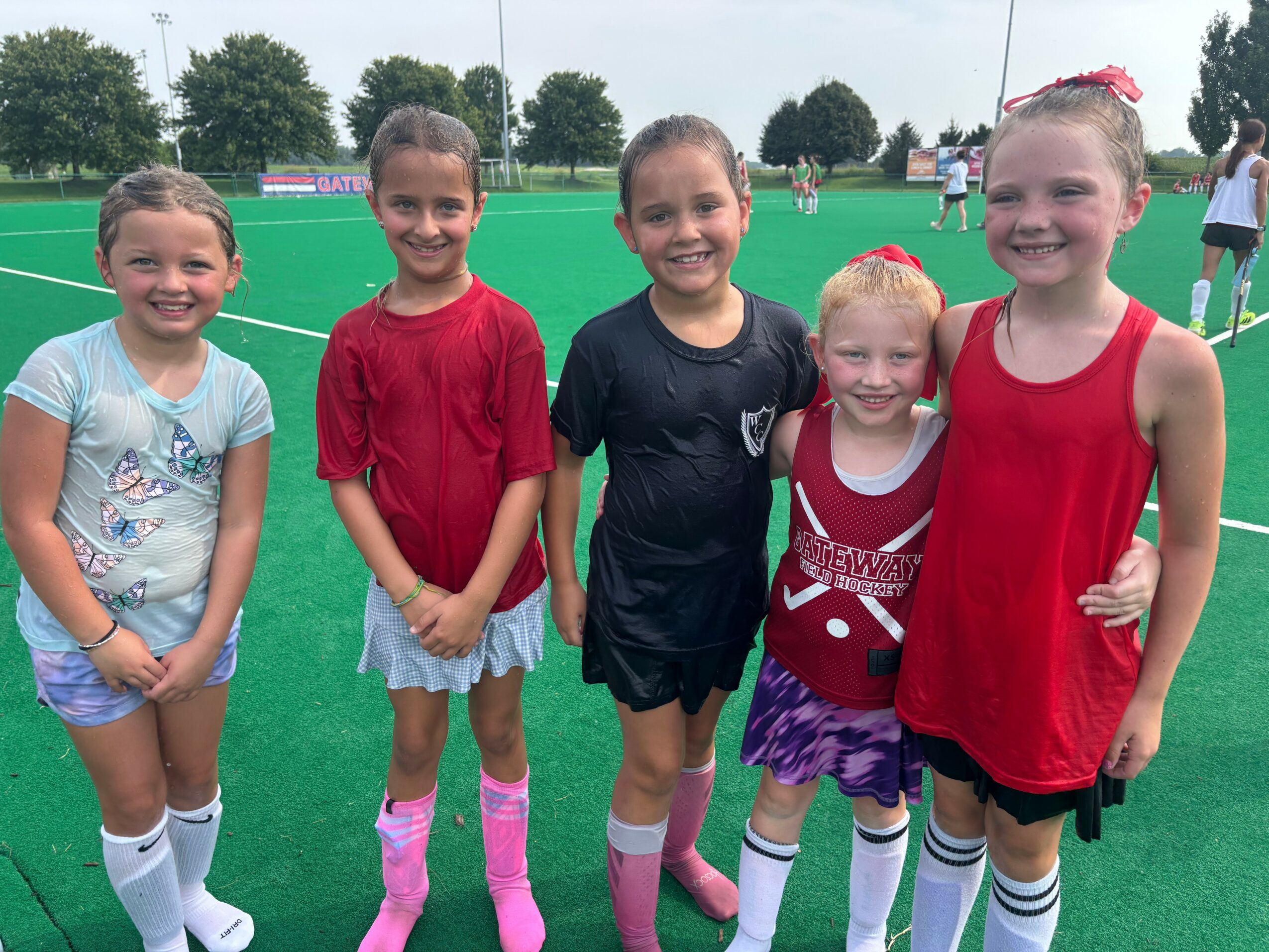 Five young girls standing on a sports field, smiling, wearing colorful sports outfits and socks, with trees and people in the background.