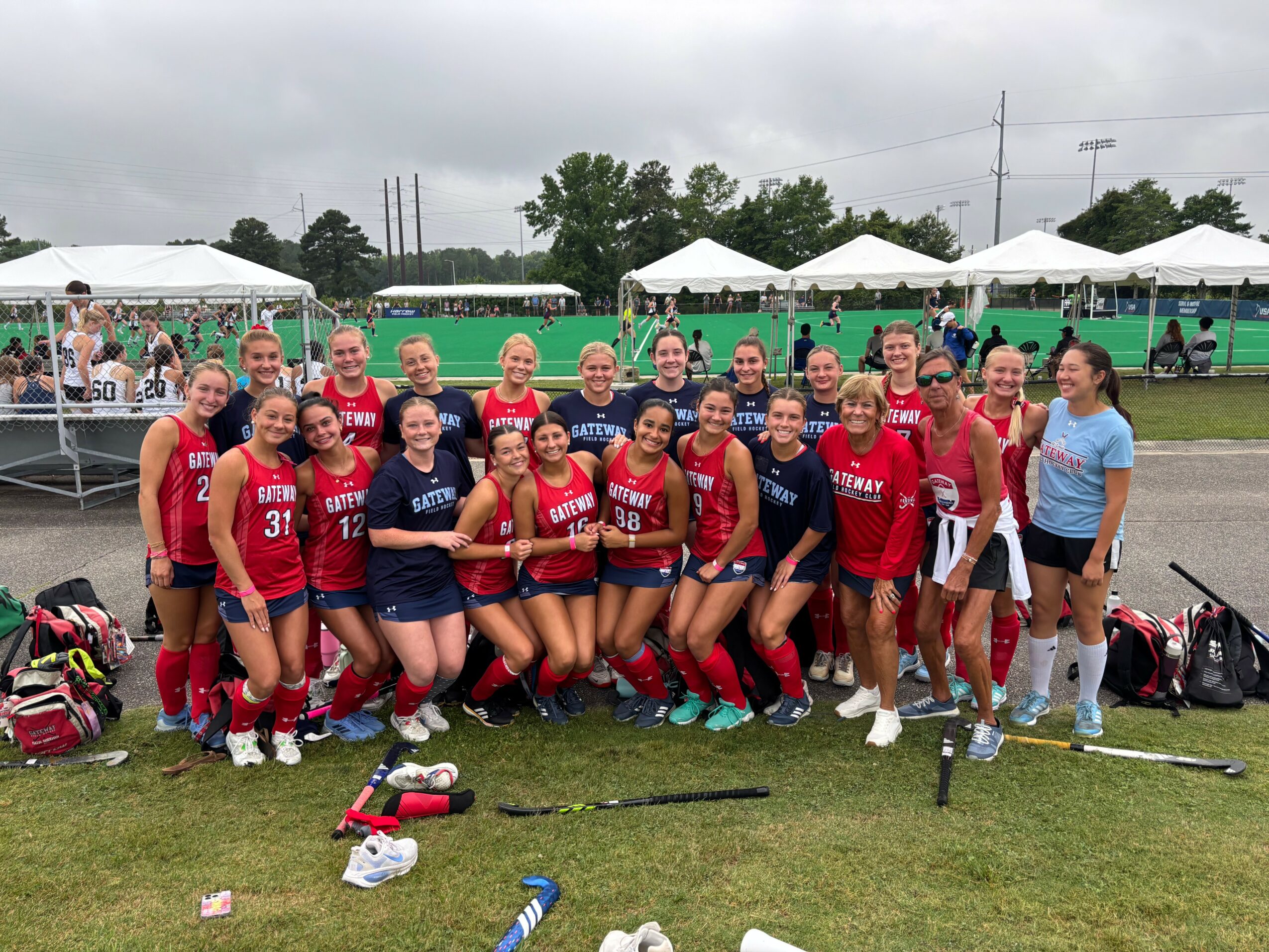 Group of female athletes in red and blue uniforms posing outdoors on grass, with tents and spectators in background.