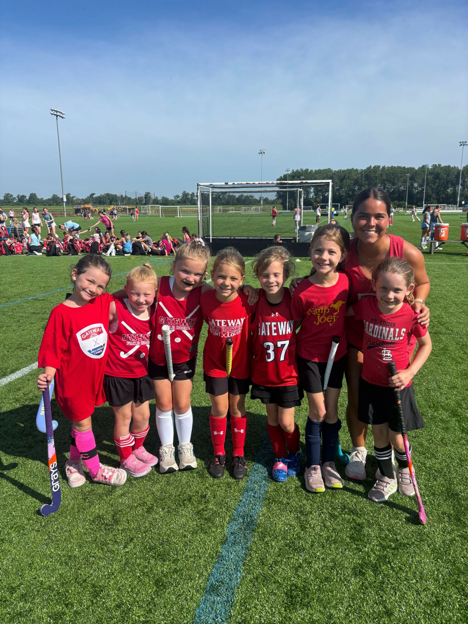 Group of young girls in red sports uniforms standing on a grassy field, with a soccer goal and other players in the background.