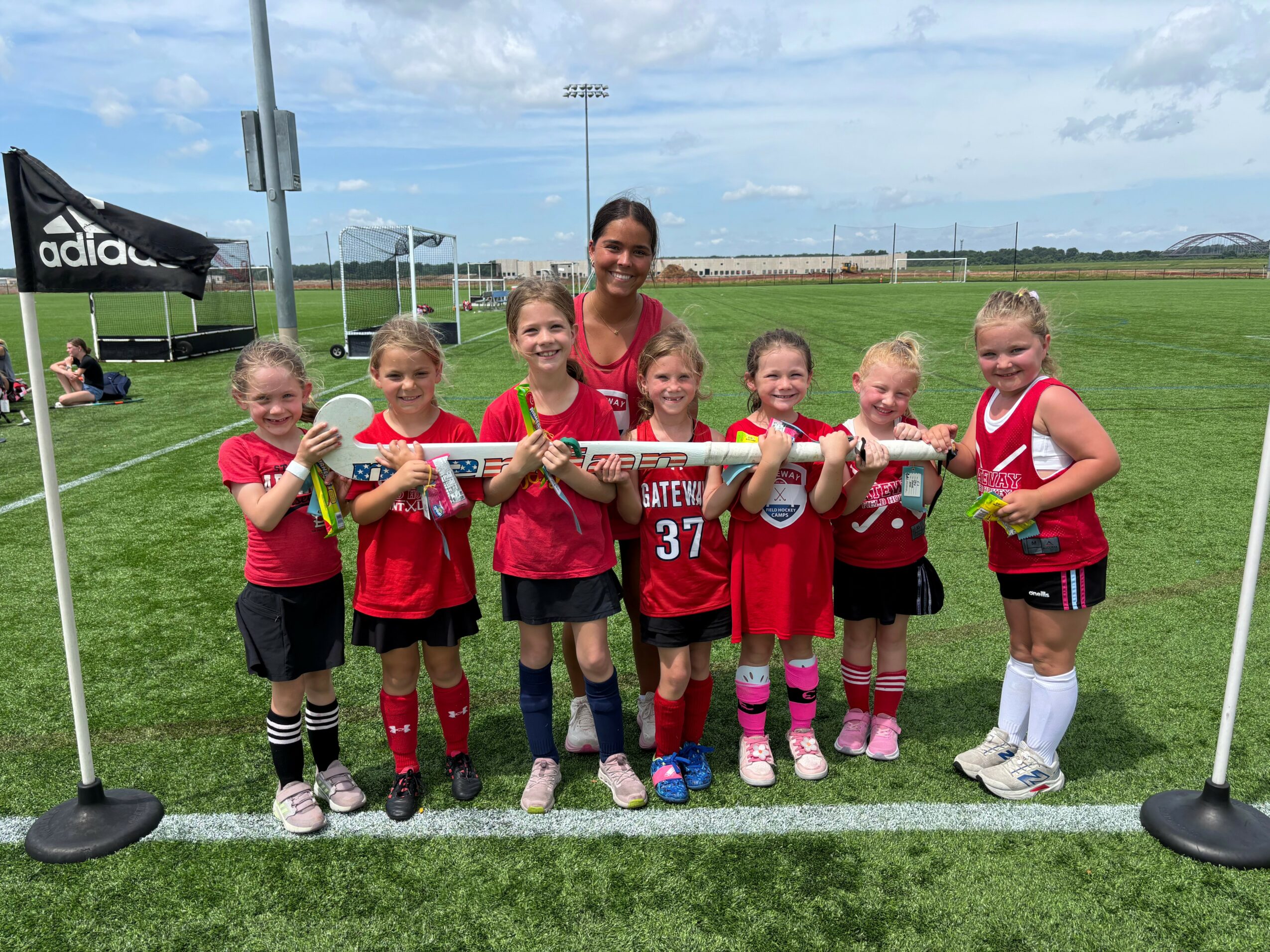 Group of young girls and a woman standing on a sports field, holding a relay baton, smiling, with a blue sky and field in background.