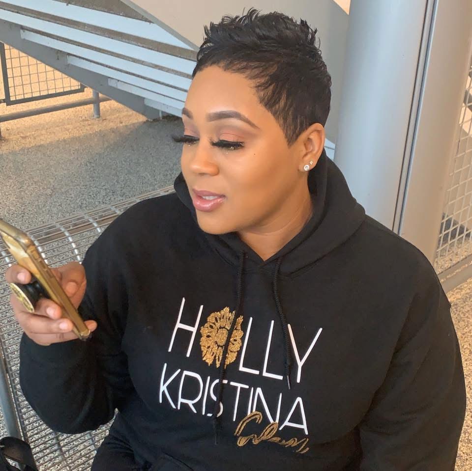 Woman with short hair wearing a black hoodie with text, looking at her phone, sitting outdoors near a metal structure.