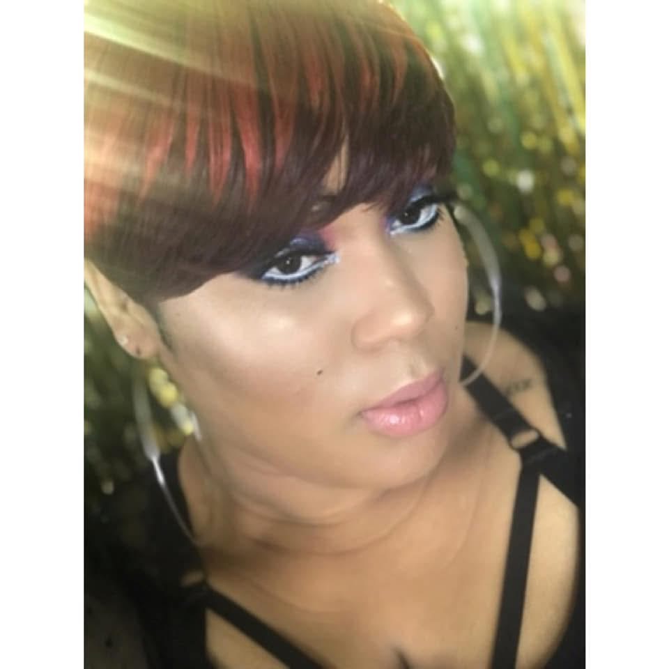 Close-up of a woman with short hair, makeup, and earrings, outdoors with sunlight and greenery in background.