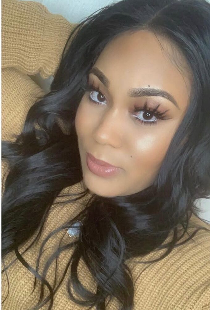 Close-up of a woman with long, wavy black hair and makeup, wearing a tan sweater, looking at the camera.