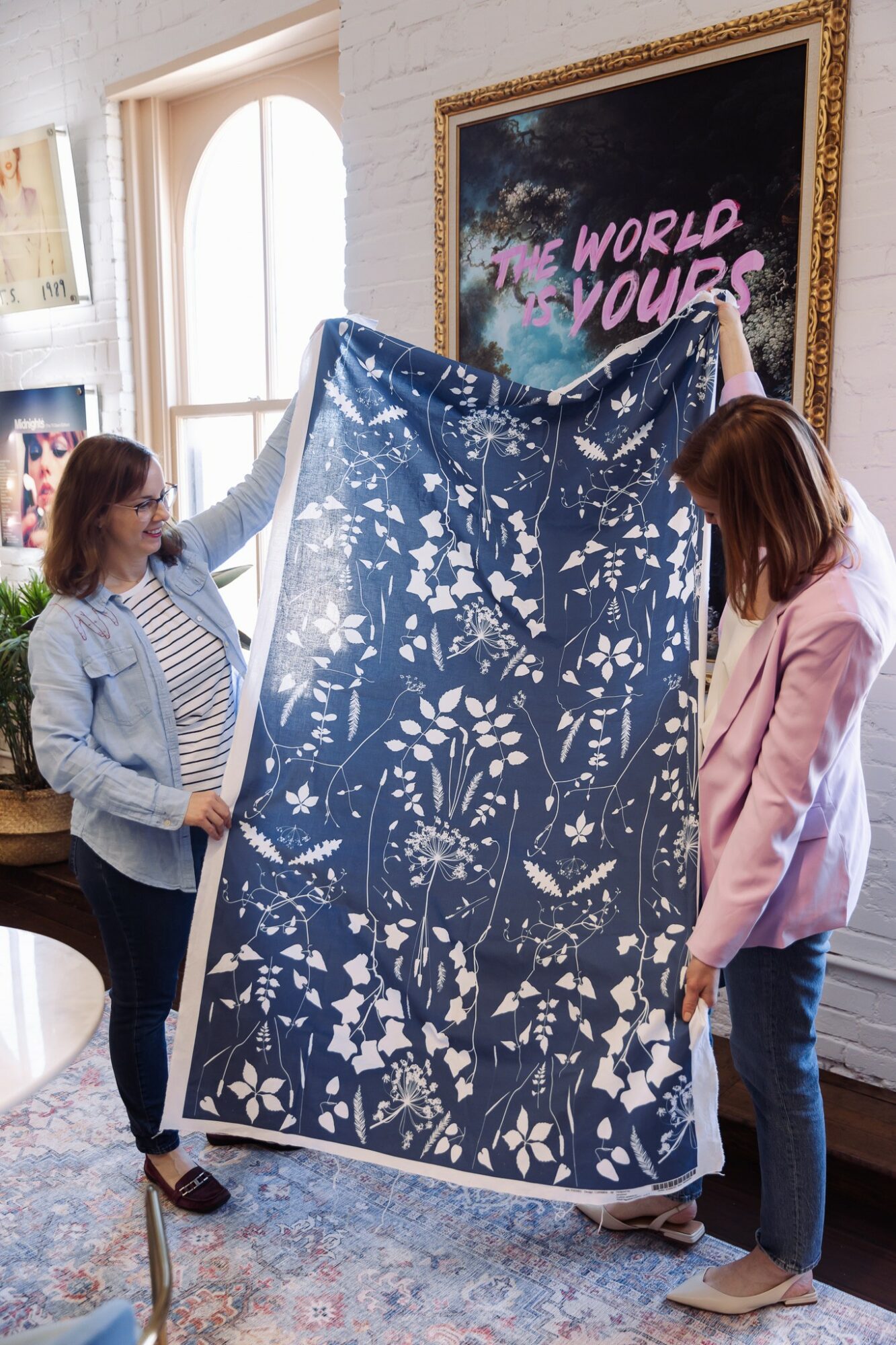 Two women hold a large blue fabric with white floral patterns indoors, near a framed sign that says 'The World is Yours'.
