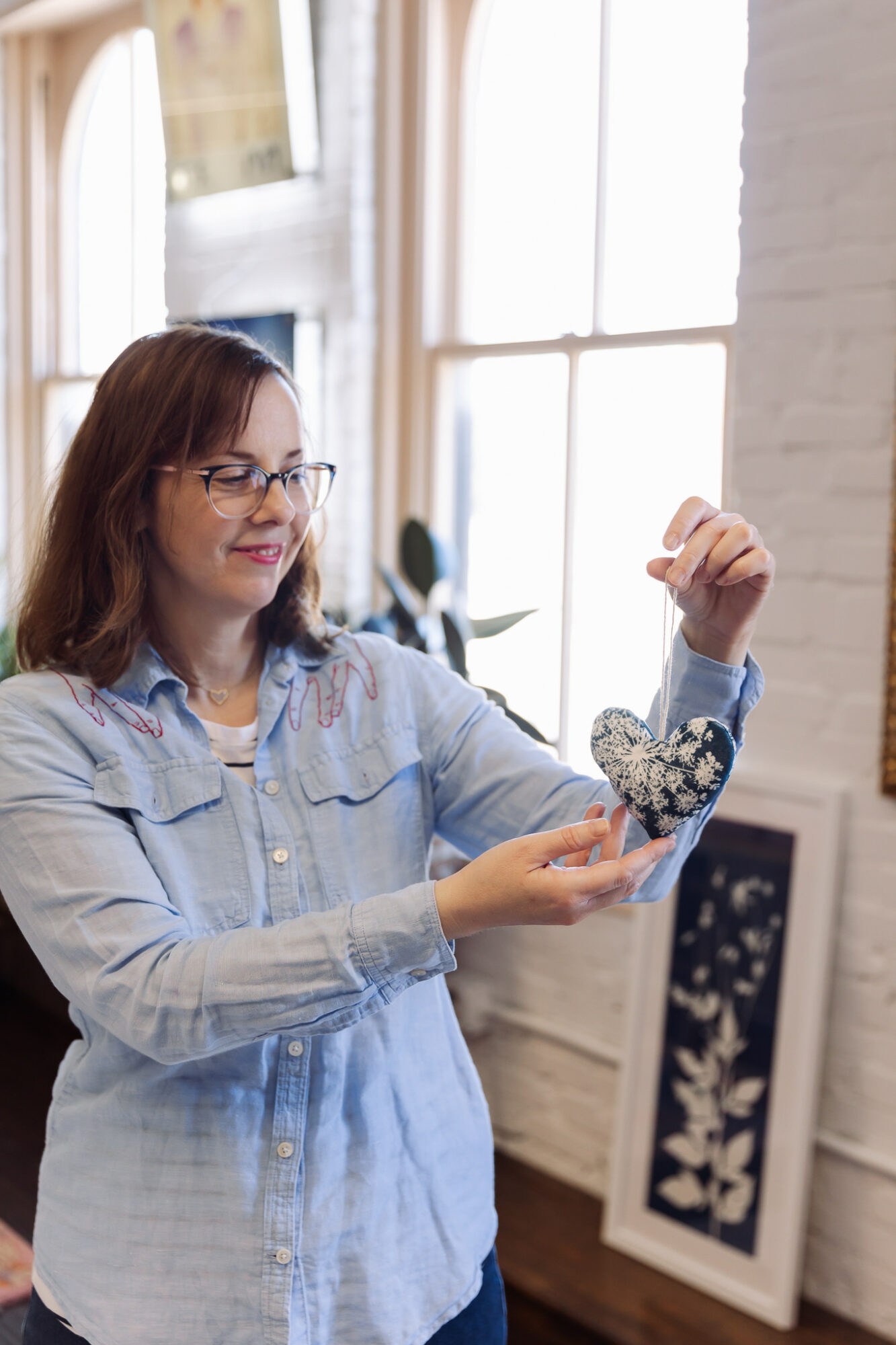 Woman with glasses holds a heart-shaped object decorated with flowers indoors near a window.