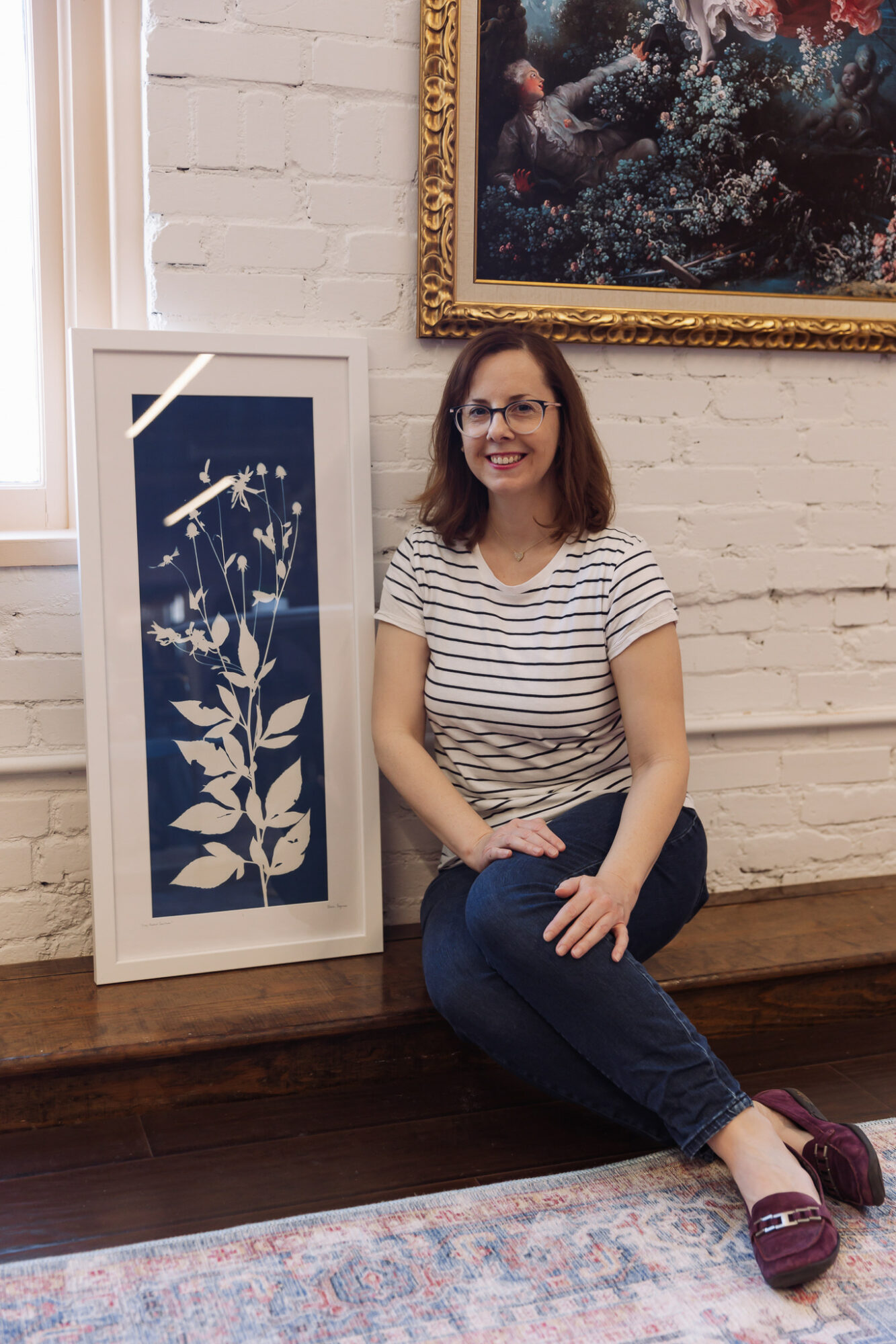 Woman with glasses, striped shirt, and jeans sitting on a wooden bench in an art gallery, smiling, with framed artwork behind her.