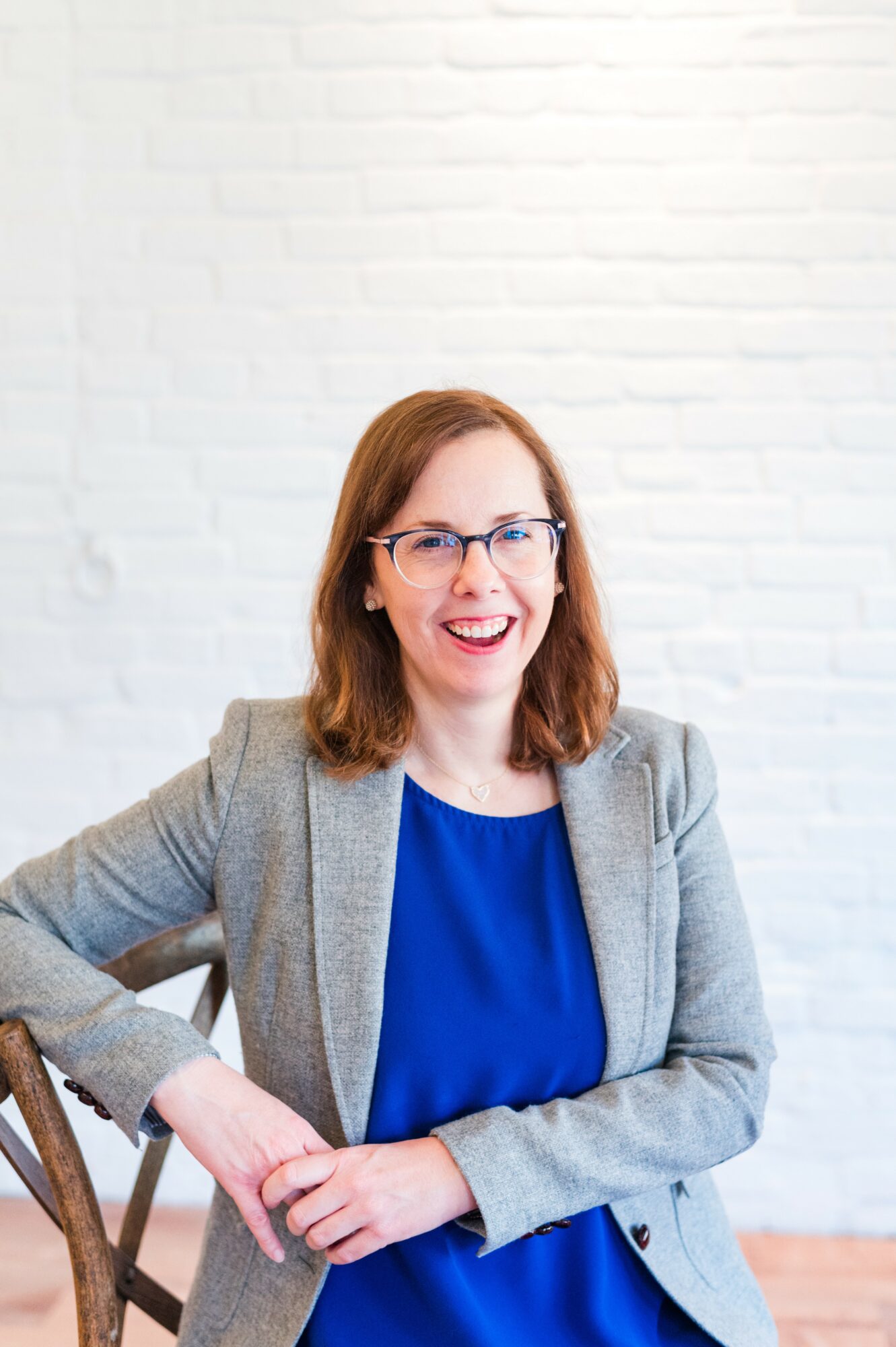 Woman with glasses and red hair smiling, wearing a gray blazer and blue top, sitting against a white brick wall.