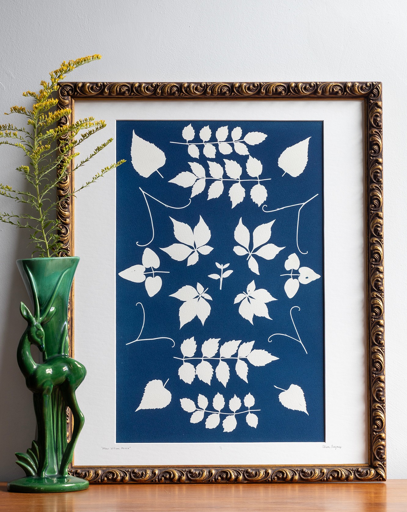 Decorative framed artwork with white leaf patterns on a blue background, next to a green vase with yellow flowers.