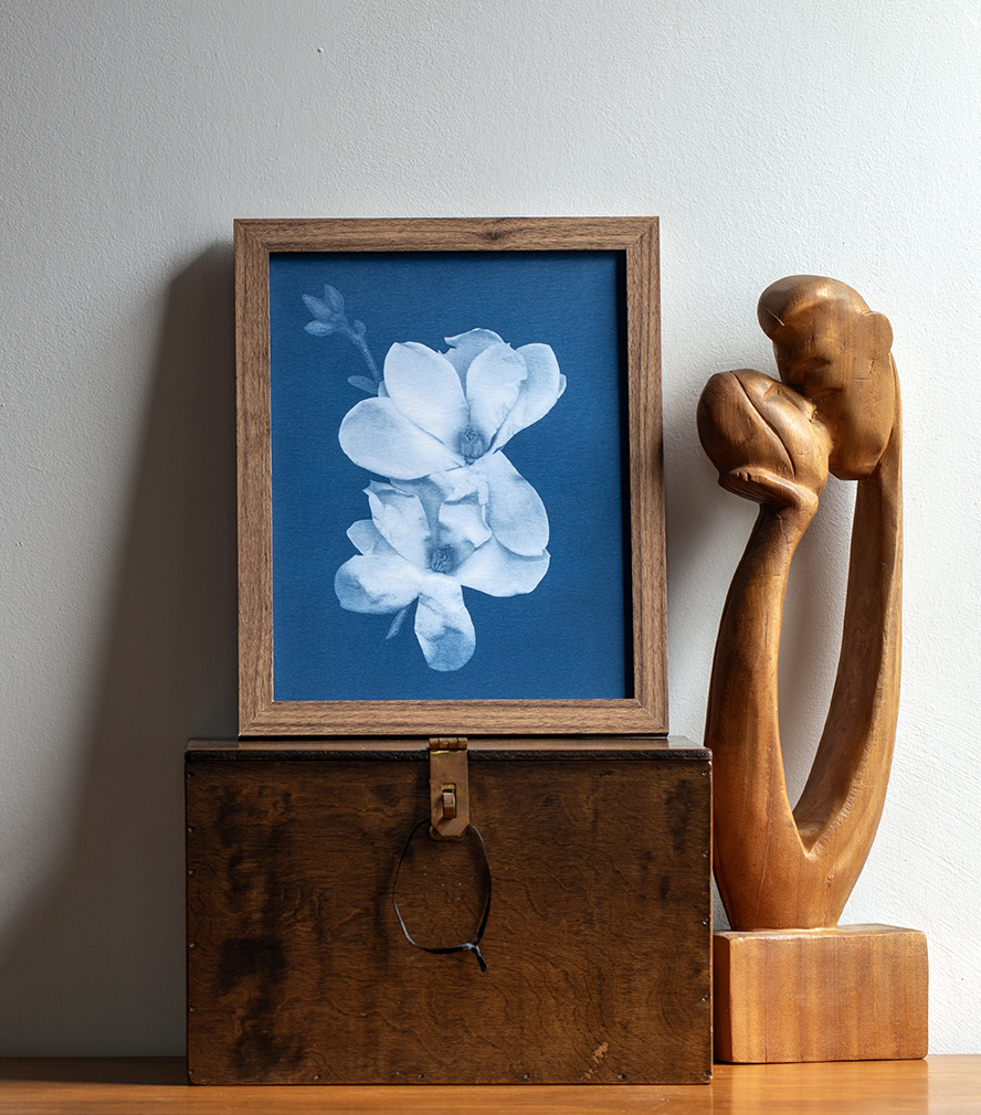 Framed flower artwork, wooden box with a ring, and a wooden sculpture of two intertwined hands, all on a light wall.