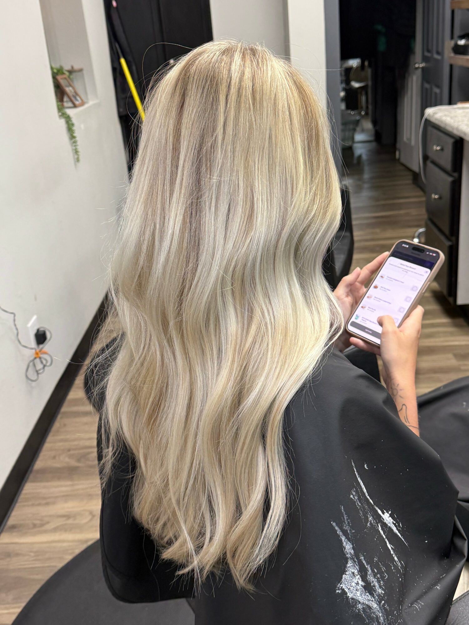 Person with long, wavy blonde hair looking at a smartphone in a salon chair.