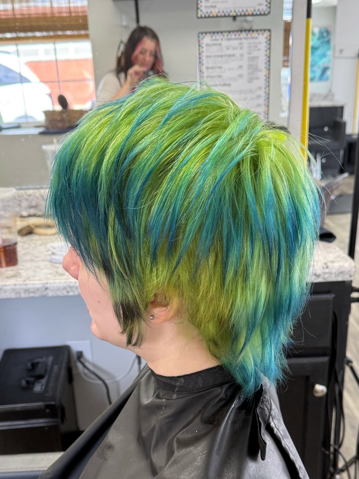 Person with short, layered hair dyed in shades of green, blue, and yellow, inside a salon. Background shows a woman and salon tools.