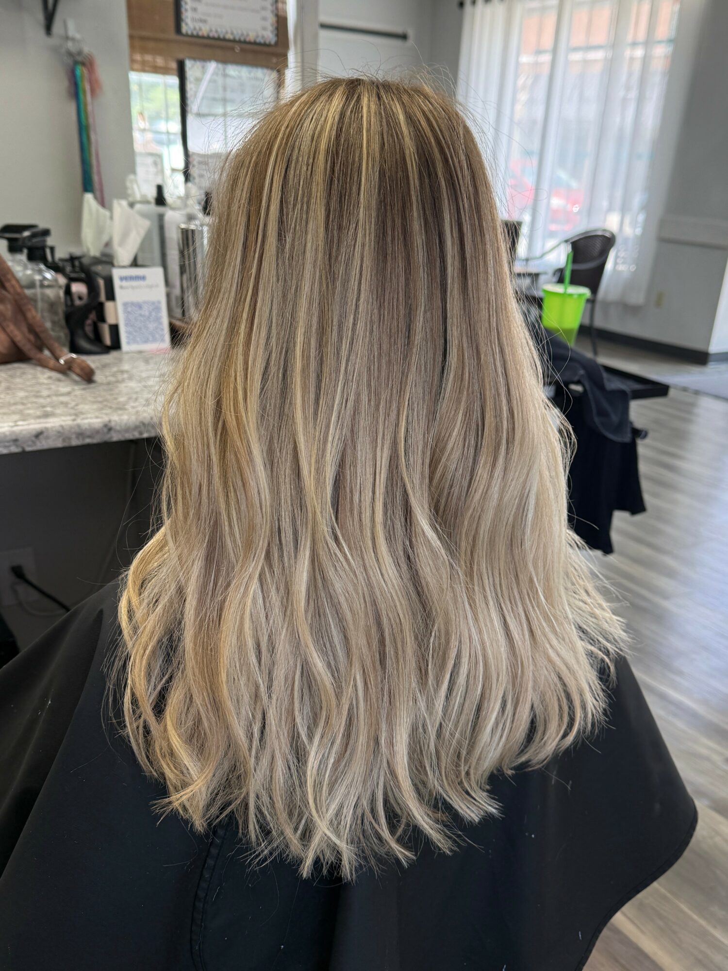 Back view of a woman with long, wavy blonde hair in a salon, with a mirror and salon tools in the background.