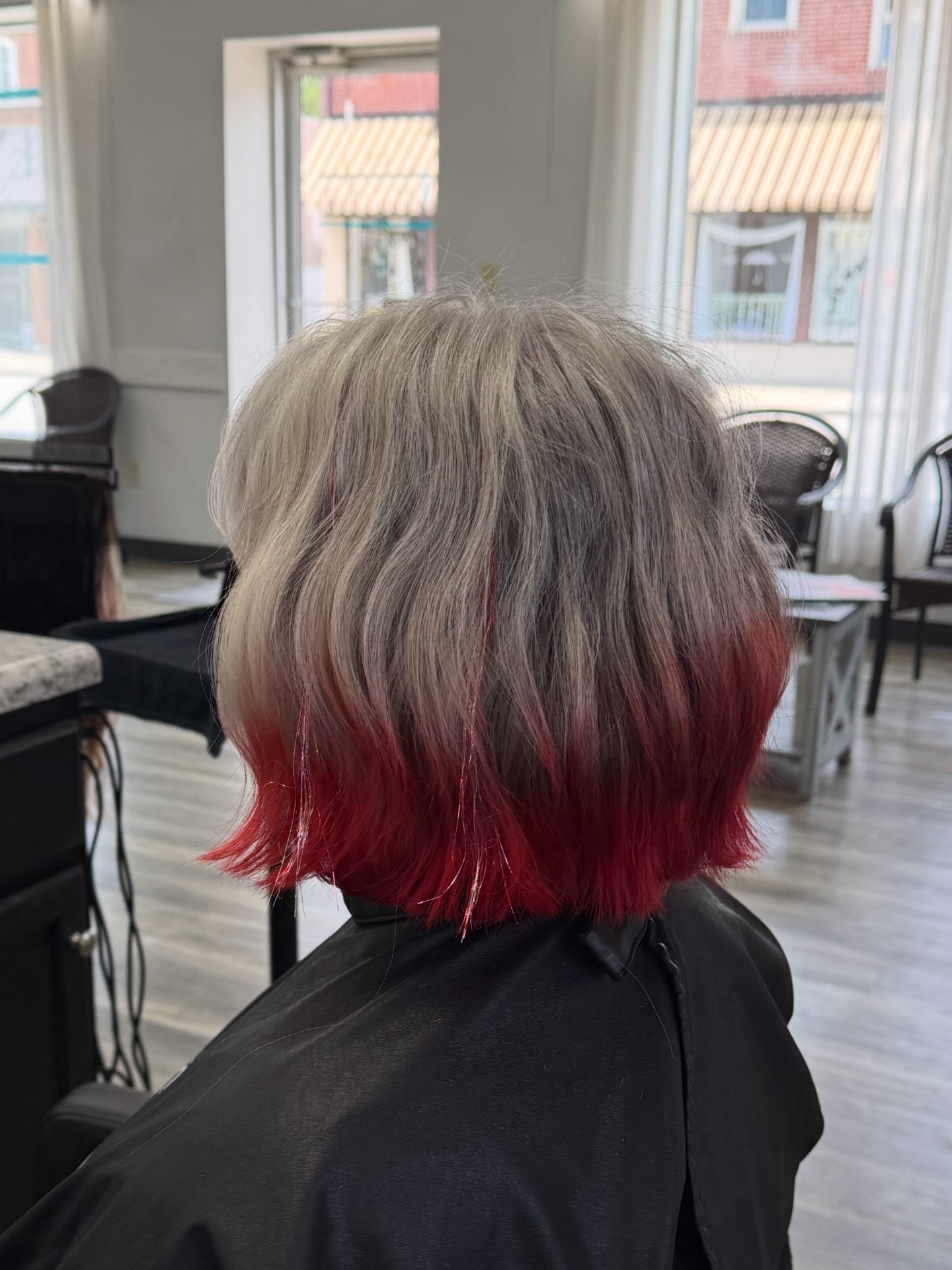 Back view of a person's short, layered hair with blonde and red colors, sitting in a salon with large windows and chairs.