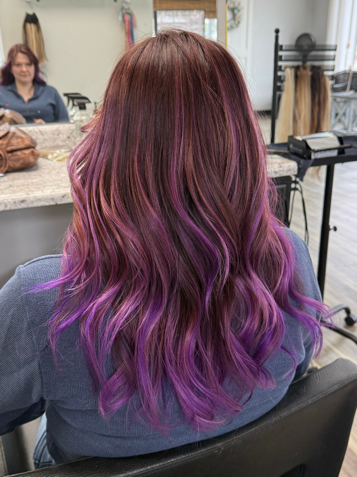 Back view of a woman with long, wavy hair dyed in a gradient from reddish-brown to purple and pink.