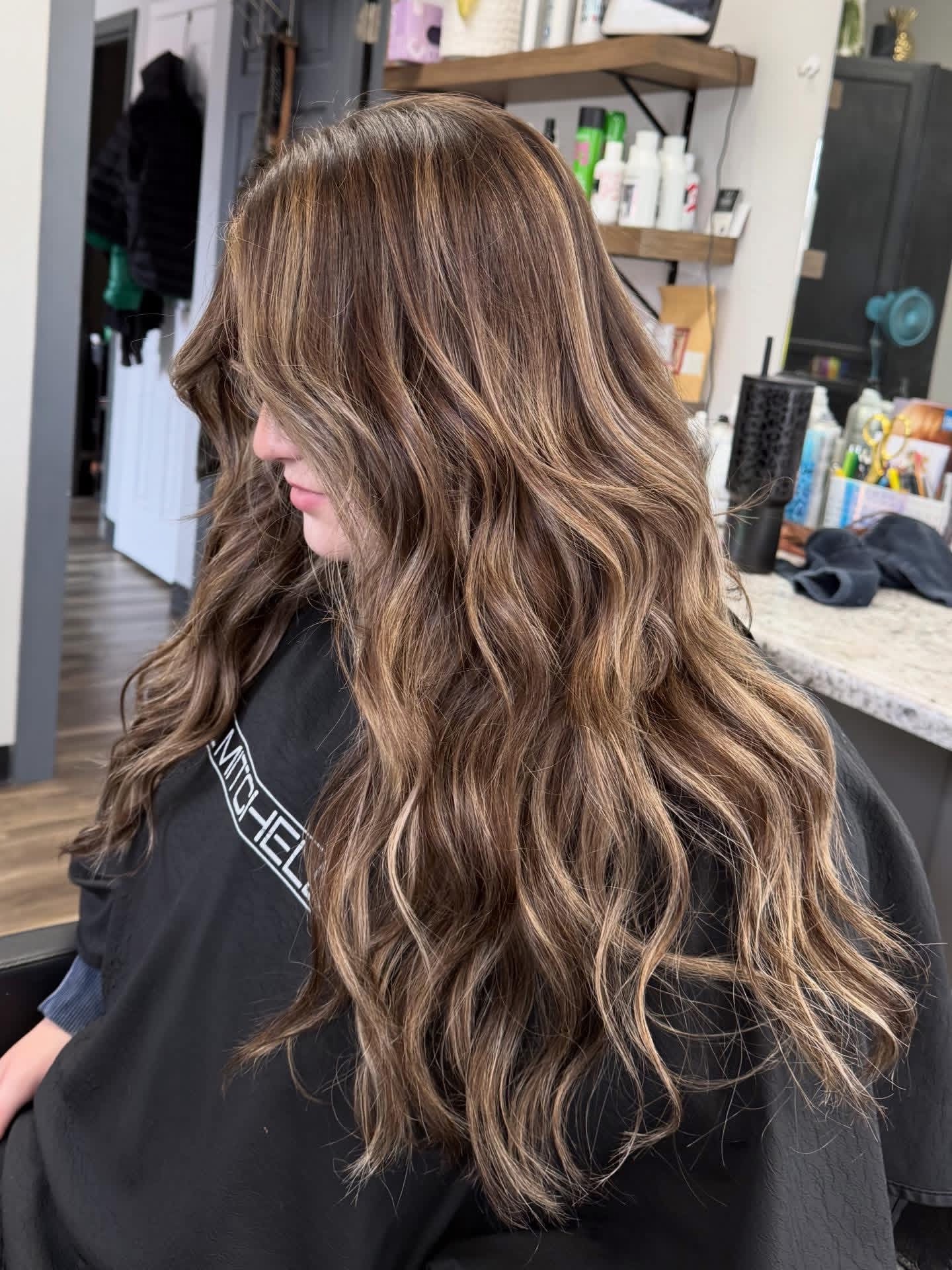 Side view of a woman with long, wavy brown hair in a salon, shelves with hair products in the background.