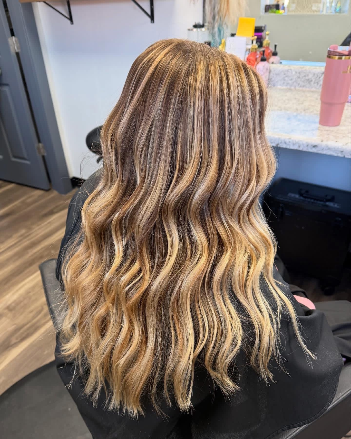 Back view of a woman with long, wavy, blonde hair in a salon, sitting at a counter with hair products.