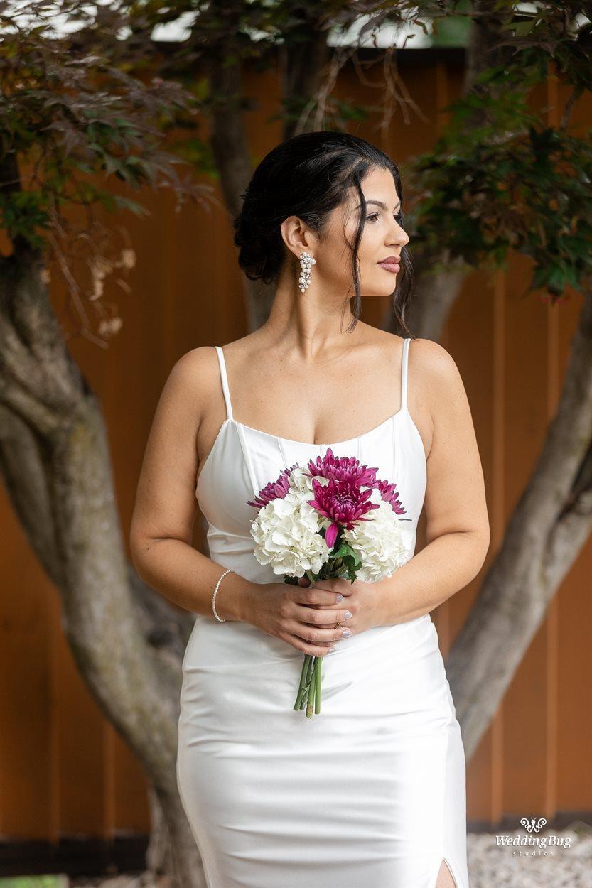 Woman in white dress holding a bouquet of purple and white flowers, standing outdoors with trees and a wooden fence behind her.