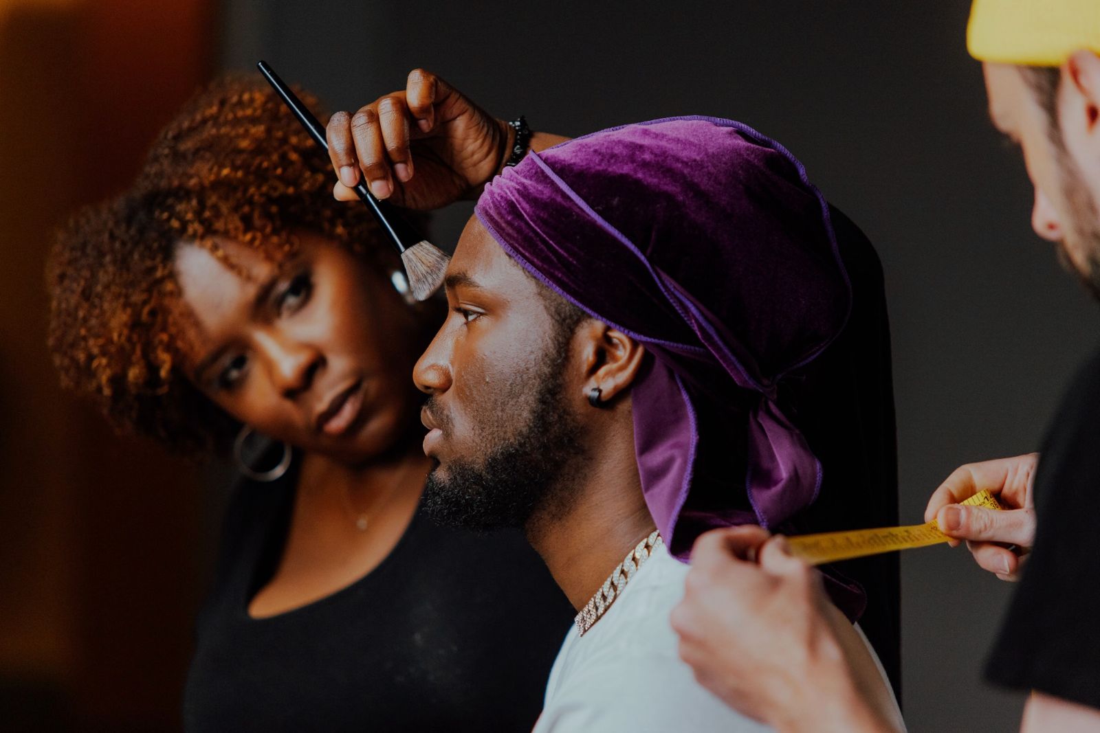 A person with a purple headscarf has makeup applied by a woman with curly hair, while another person holds a brush nearby.