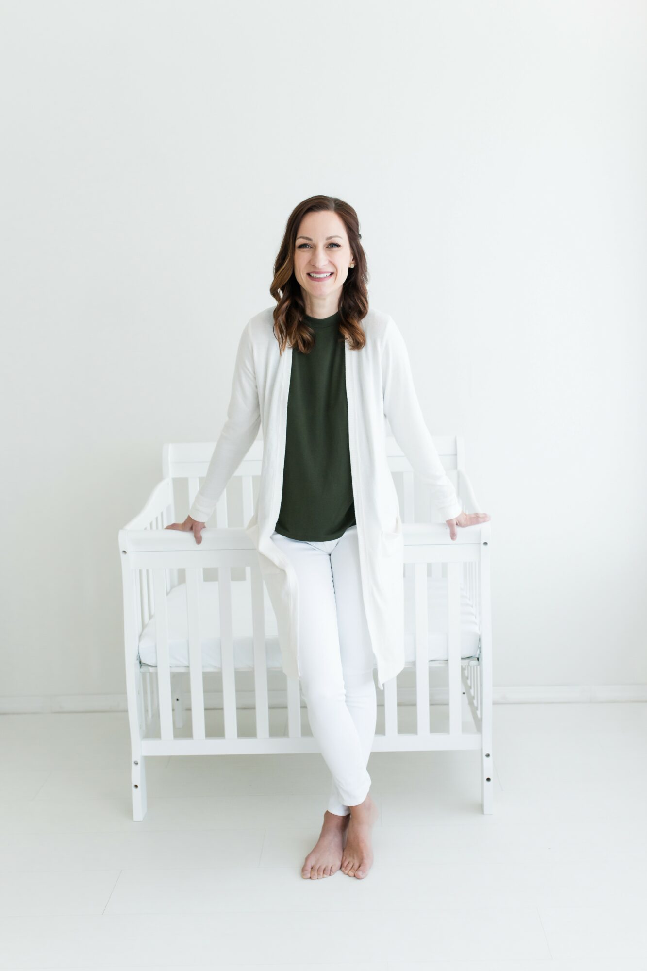 Woman standing barefoot in front of a white crib, smiling, wearing a white cardigan and dark top, against a plain white background.
