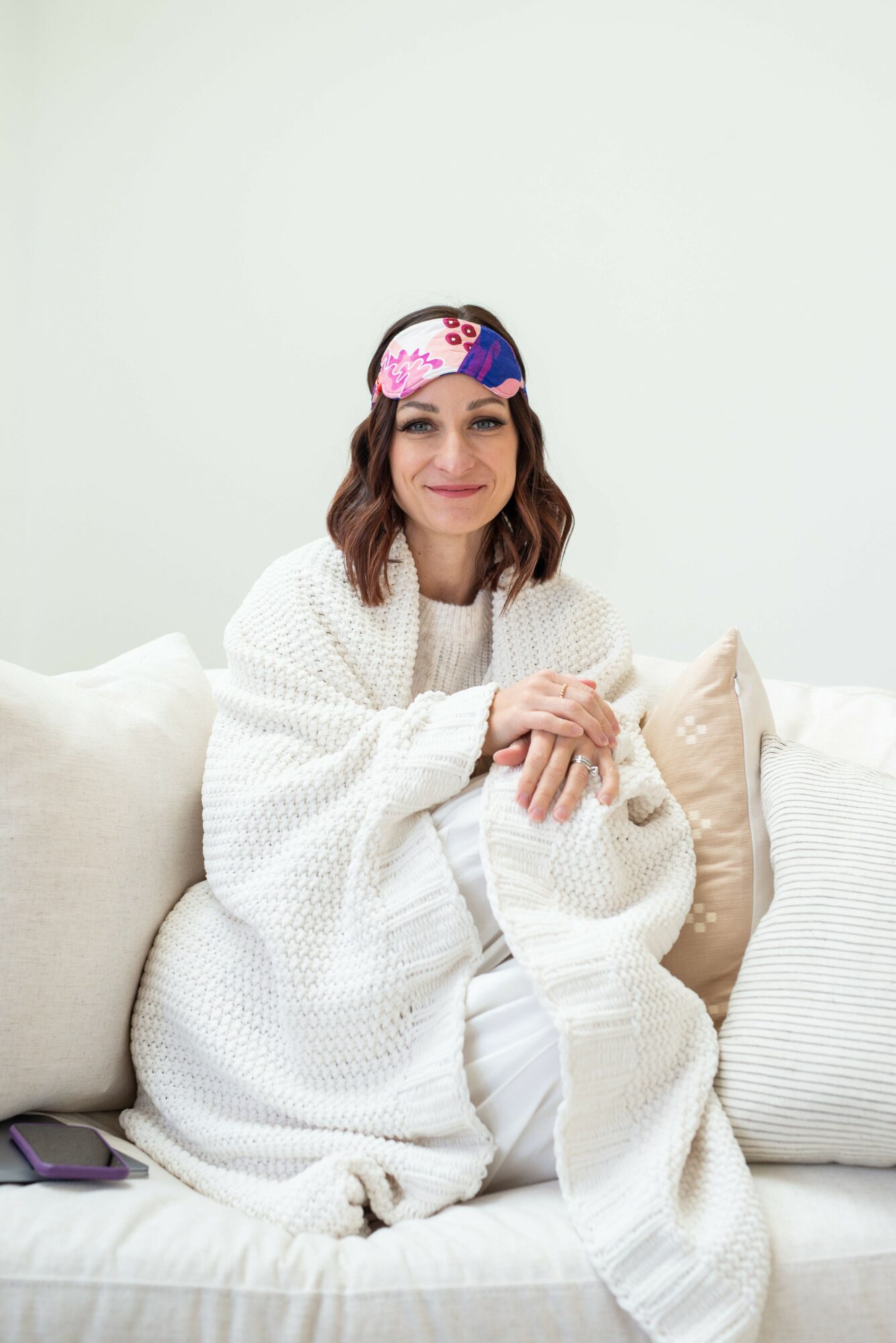 Woman sitting on a sofa wearing a colorful sleep mask, smiling, with white blanket and pillows around her.