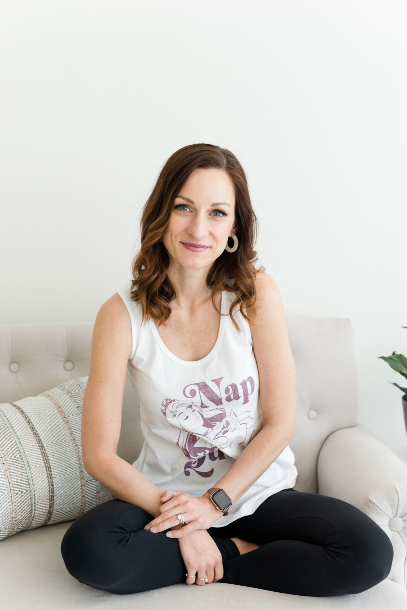Woman with shoulder-length brown hair sitting cross-legged on a sofa, wearing a white tank top and black pants.