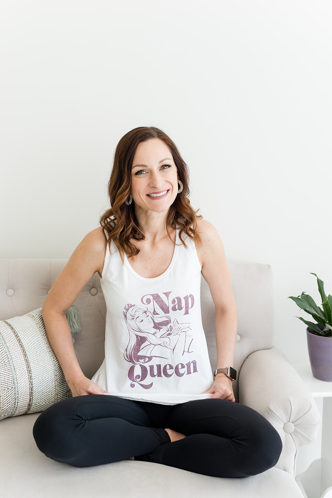 Woman sitting cross-legged on a sofa, smiling, wearing a white tank top and black pants, with a potted plant nearby.