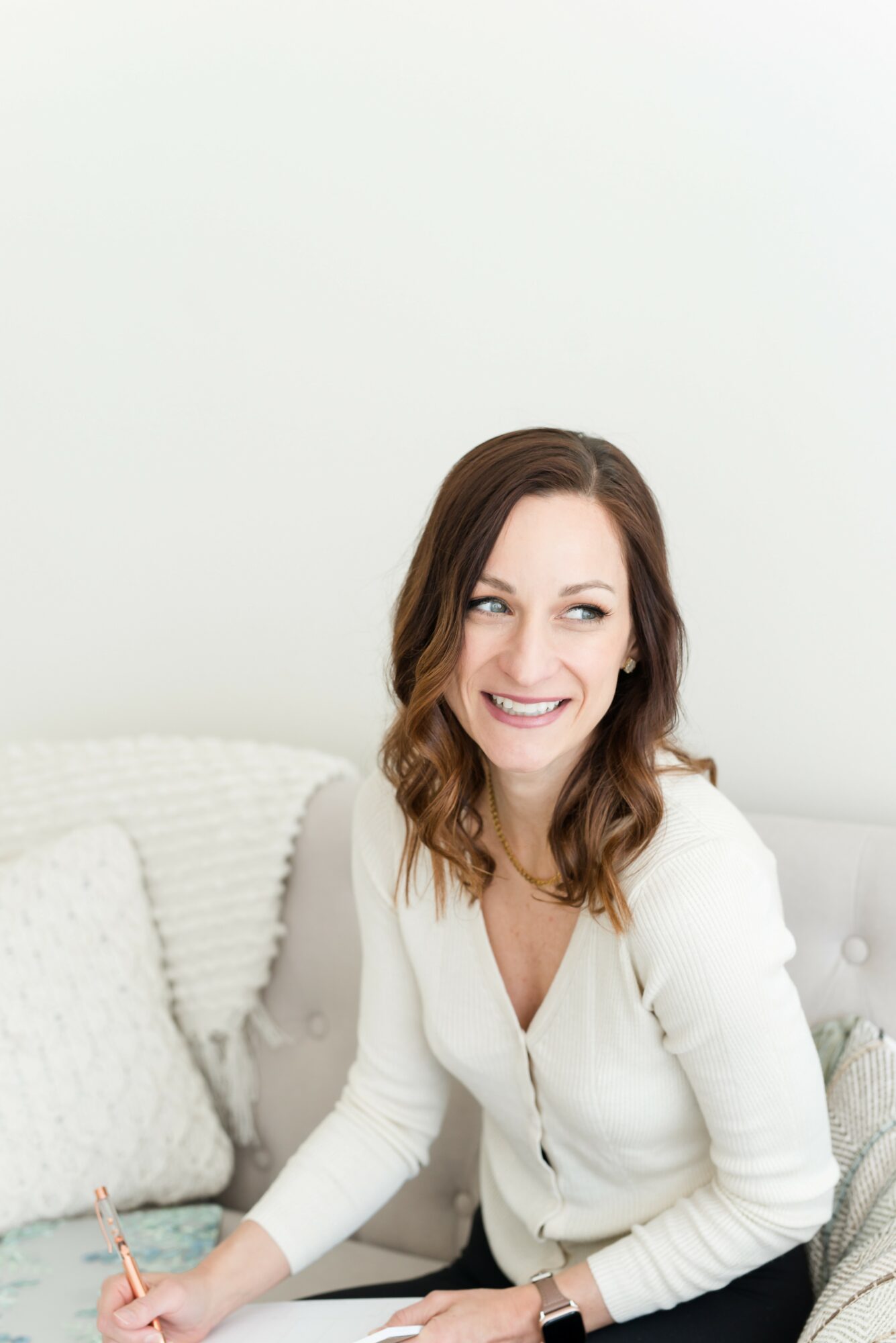 Smiling woman with wavy brown hair sitting on a white couch, wearing a white top, holding a pen, in a bright room.