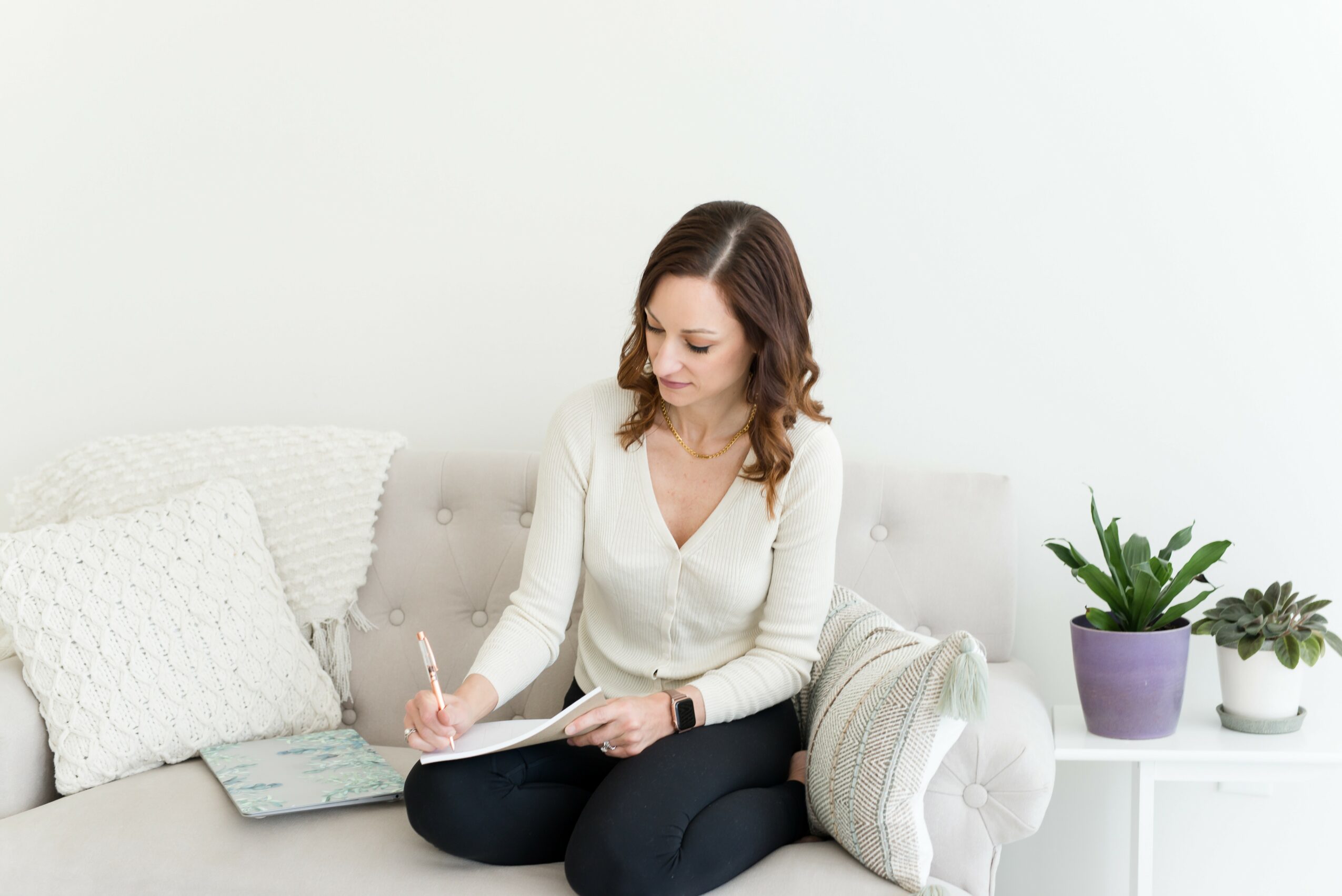 Woman sitting on a sofa, writing in a notebook, with pillows and potted plants nearby.