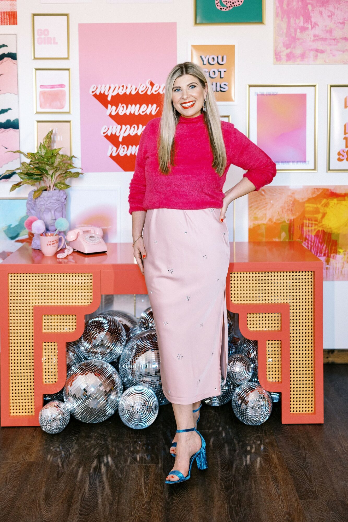 Woman in pink sweater and skirt standing in front of colorful wall with decorations and disco balls.