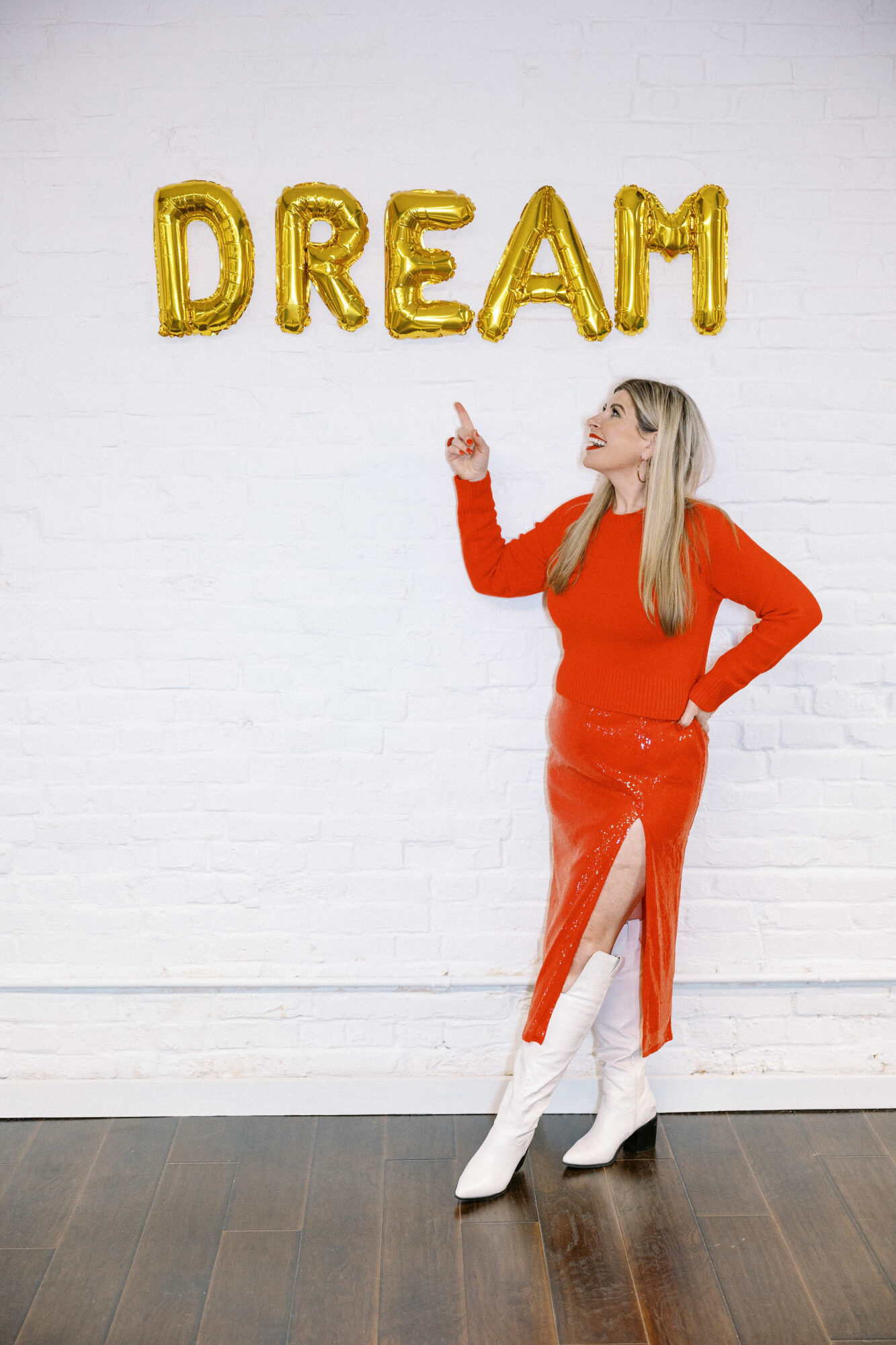 Woman in red dress and white boots pointing upward, gold balloons spelling 'DREAM' on white wall, wooden floor.