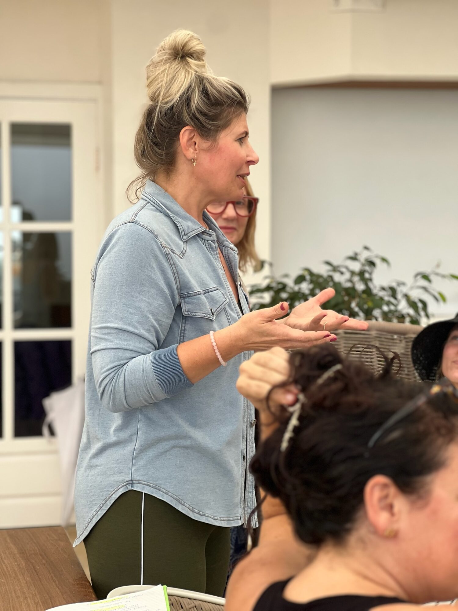 Woman with blonde hair in a bun speaking to a group indoors, with people seated around her and a plant in the background.