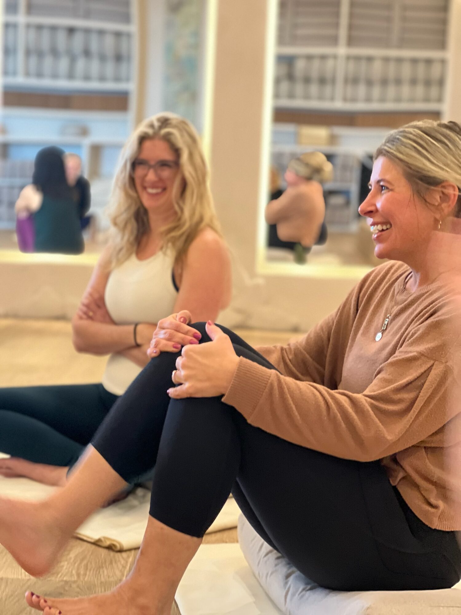 Two women sitting on a couch, smiling and engaging in conversation, with large windows behind them.