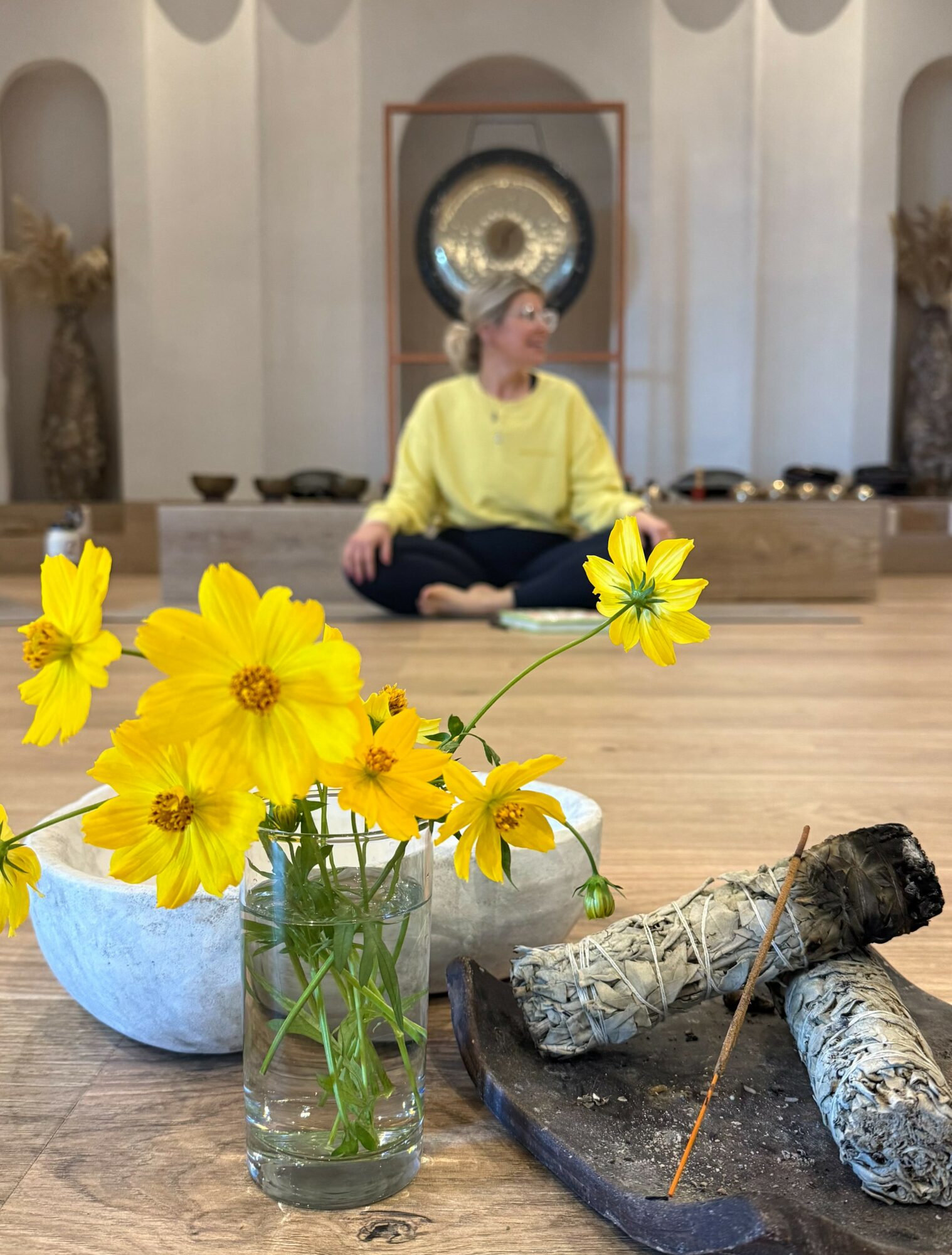 Person sitting cross-legged on floor in a room with a gong behind, yellow flowers in a glass jar in foreground, logs on a tray.