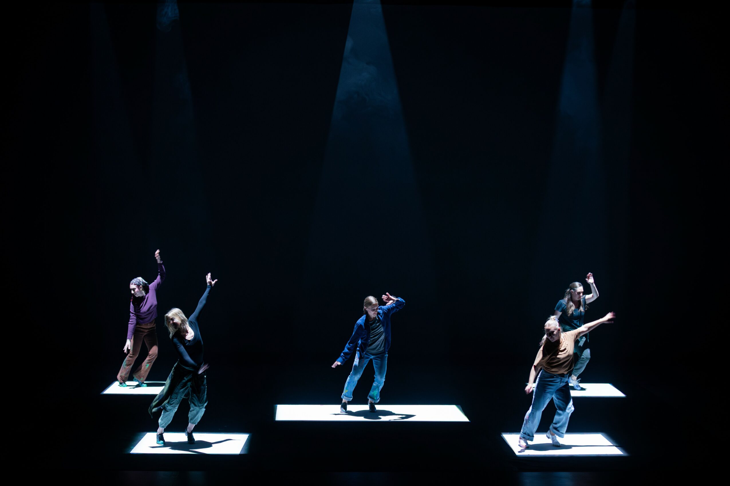 Six dancers perform on illuminated platforms in a dark stage with overhead lighting, striking various poses.