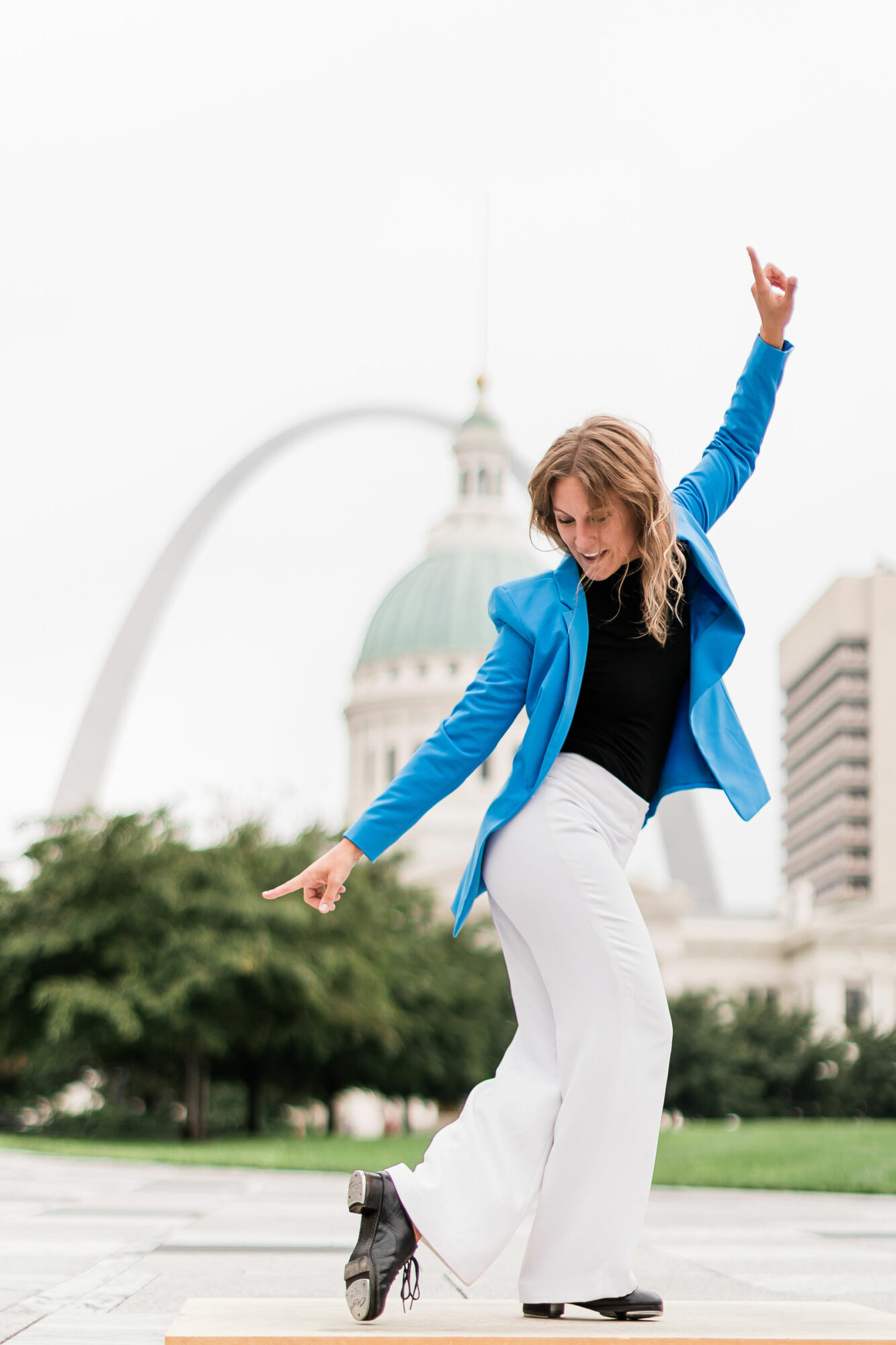 Woman dancing outdoors with cityscape and a large arch in the background, wearing a blue jacket and white pants.