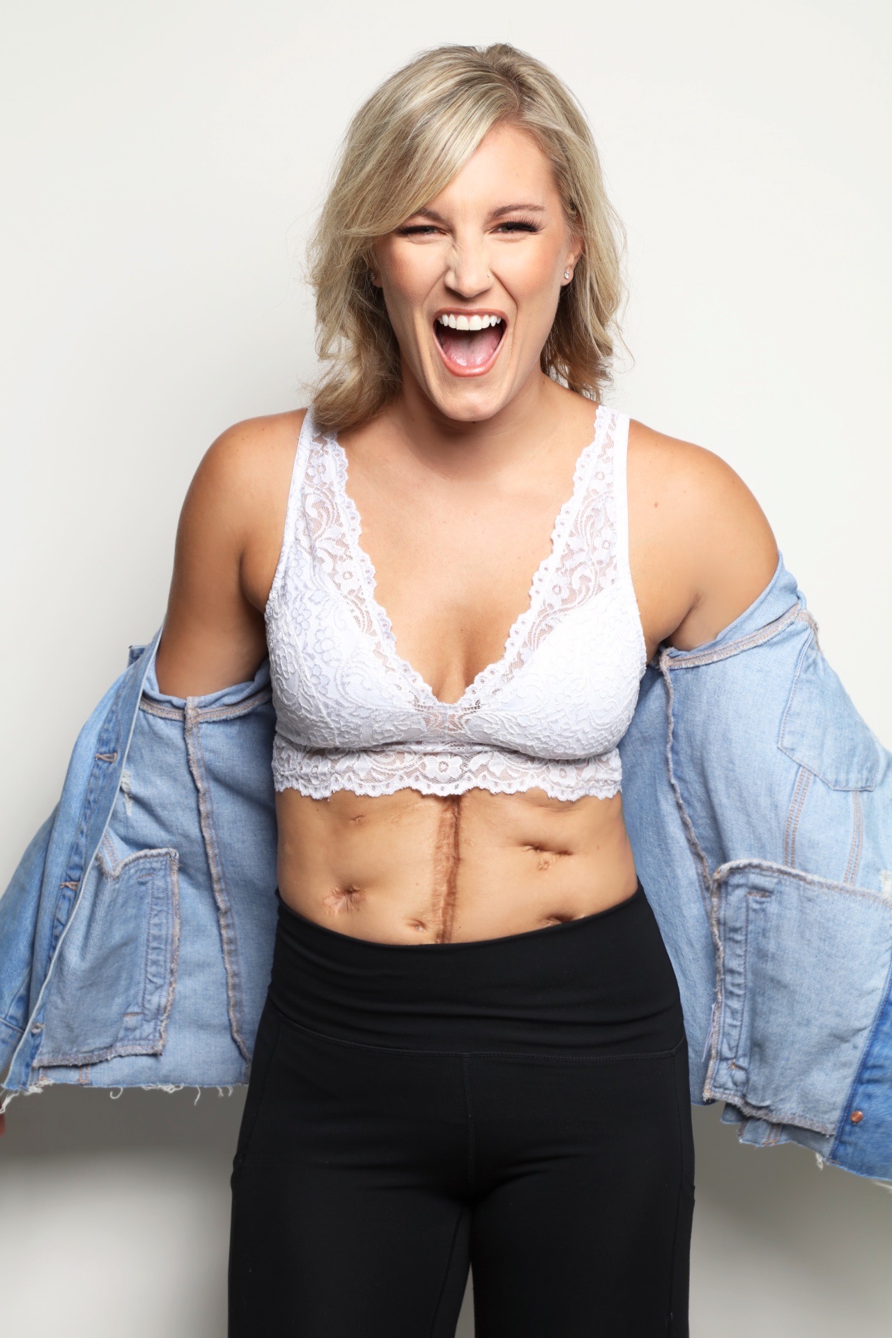 Woman with short blonde hair laughing, wearing a white lace top and black pants, holding a denim jacket off her shoulders.