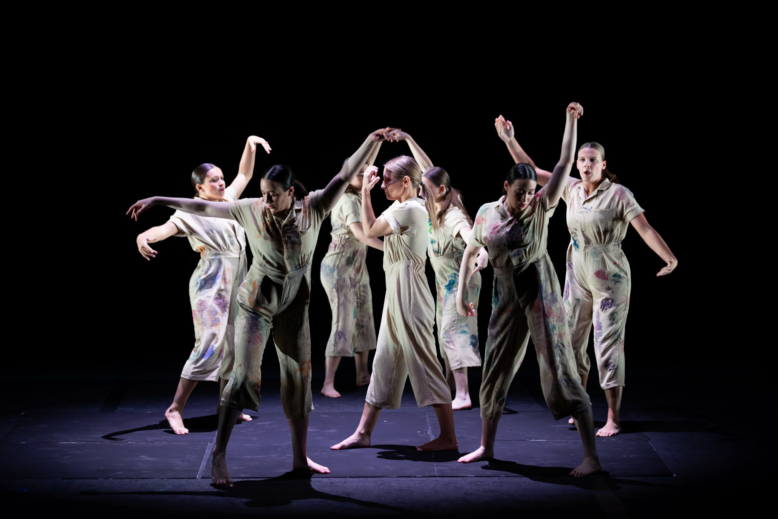 Seven dancers in flowing costumes perform on a dark stage with arms raised and extended, creating a dynamic scene.