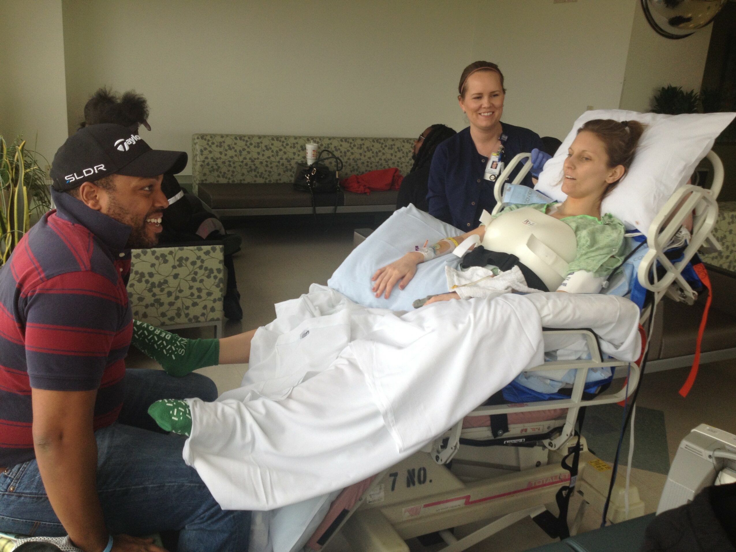 Person lying in hospital bed with bandages, surrounded by two women and a man in a hospital room.