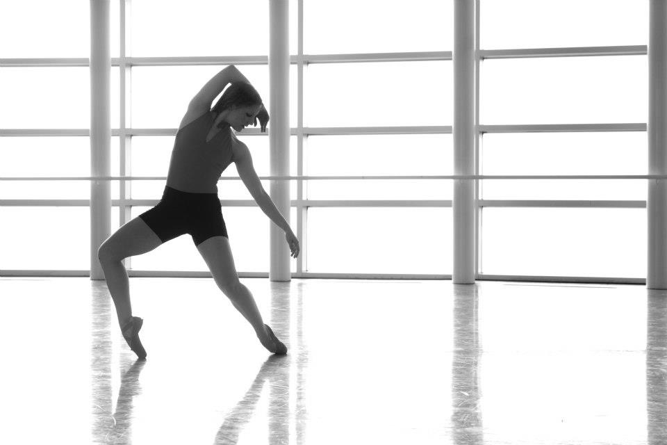 Person stretching in a dance studio with large windows, standing on one leg with the other leg extended back, arms raised.