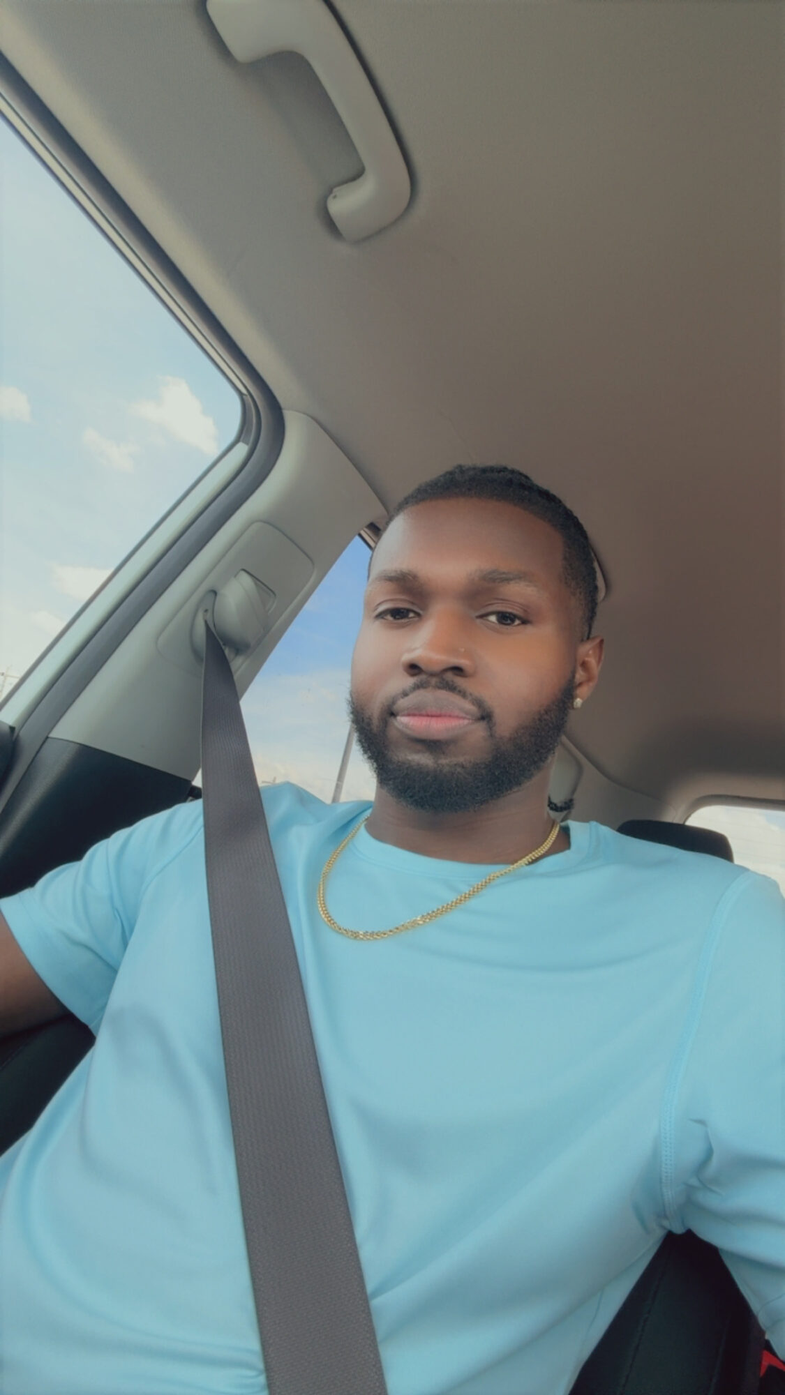 Young man with a beard and gold chain sitting in a car, wearing a light blue shirt, looking at the camera.