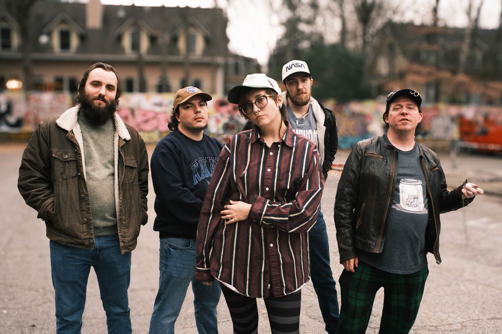 Group of five people standing outdoors in front of a building and trees, posing for a photo.